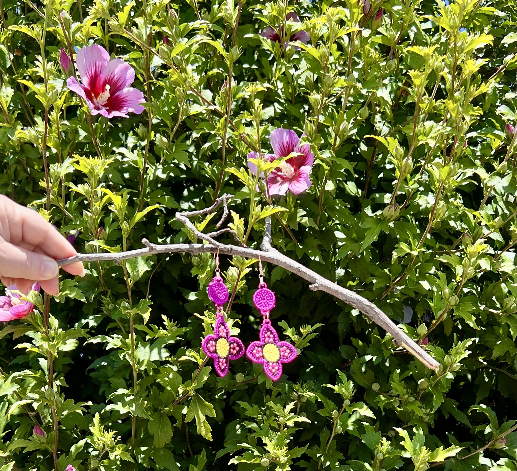 Pink beaded earrings hanging from a branch in front of green leafy bushes with pink flowers.