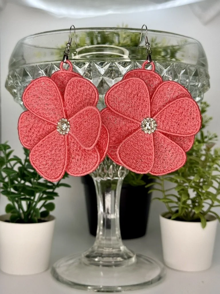Pink flower-shaped earrings with rhinestone centers hanging on a glass stand with green plants in white pots in the background.