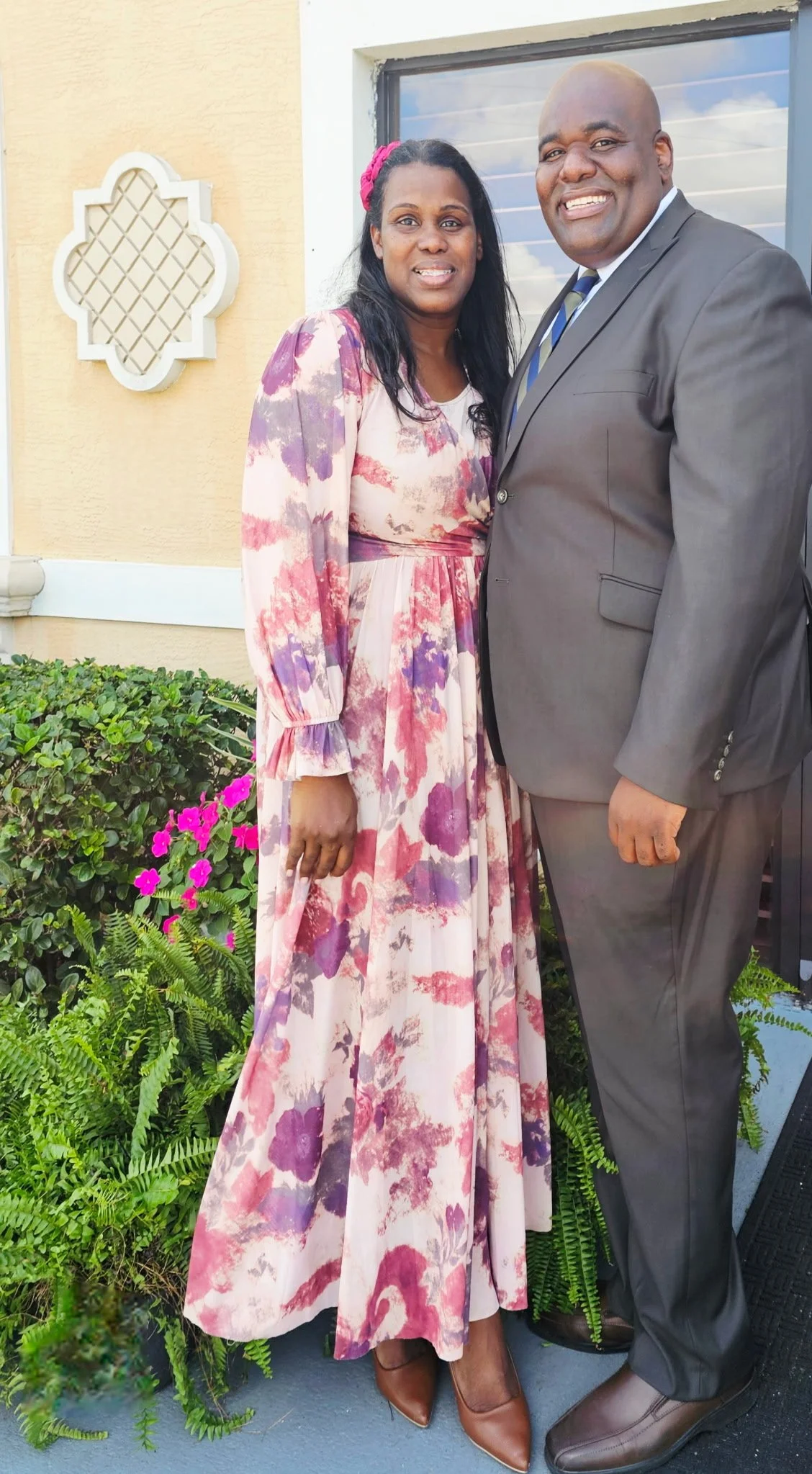 A smiling woman in a long, flowing floral dress and a man in a dark suit standing outside near a house with a window and decorative element on the wall. There are green plants and pink flowers in front of them.