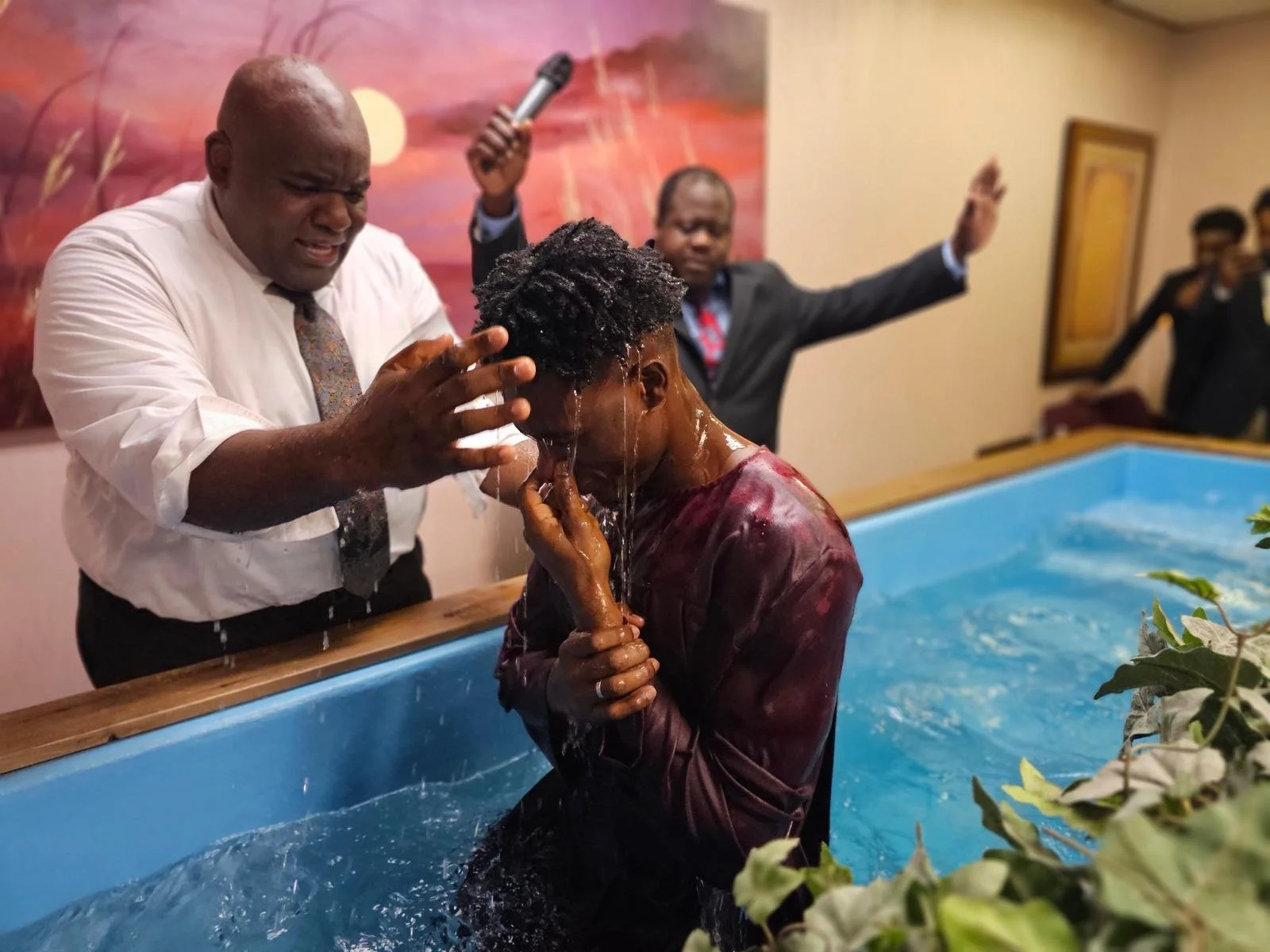 A person is being baptized in a baptismal pool by a clergyman, with water pouring over their head, and an assistant with a microphone raising their hand in the background.