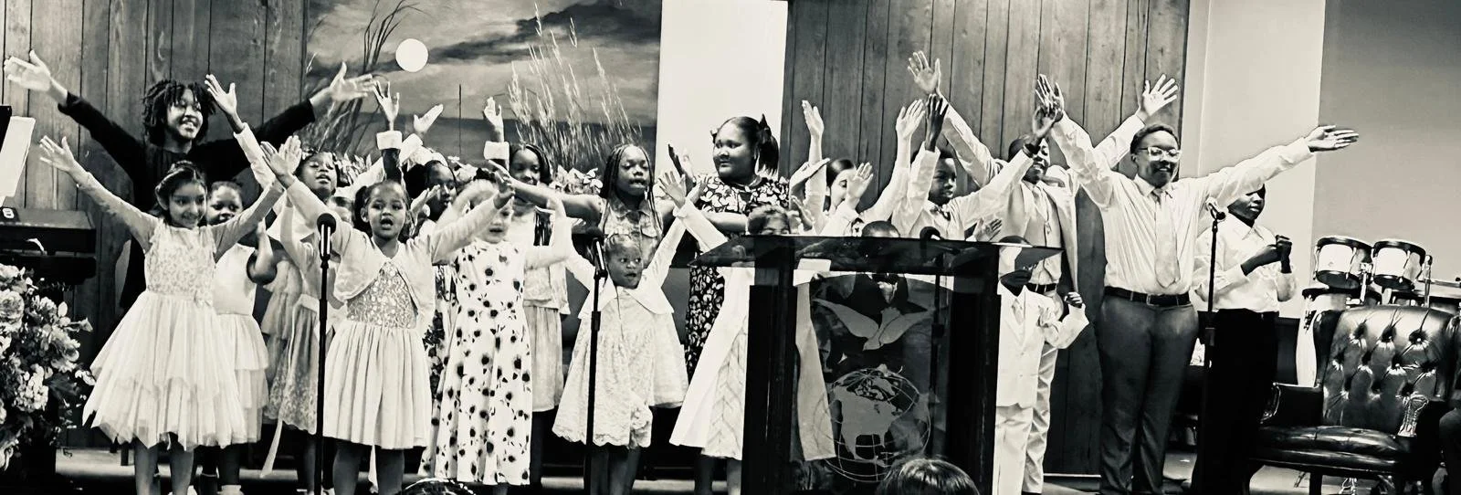 A group of children and two adults standing on stage with arms raised, in a church or auditorium setting, possibly during a performance or celebration.