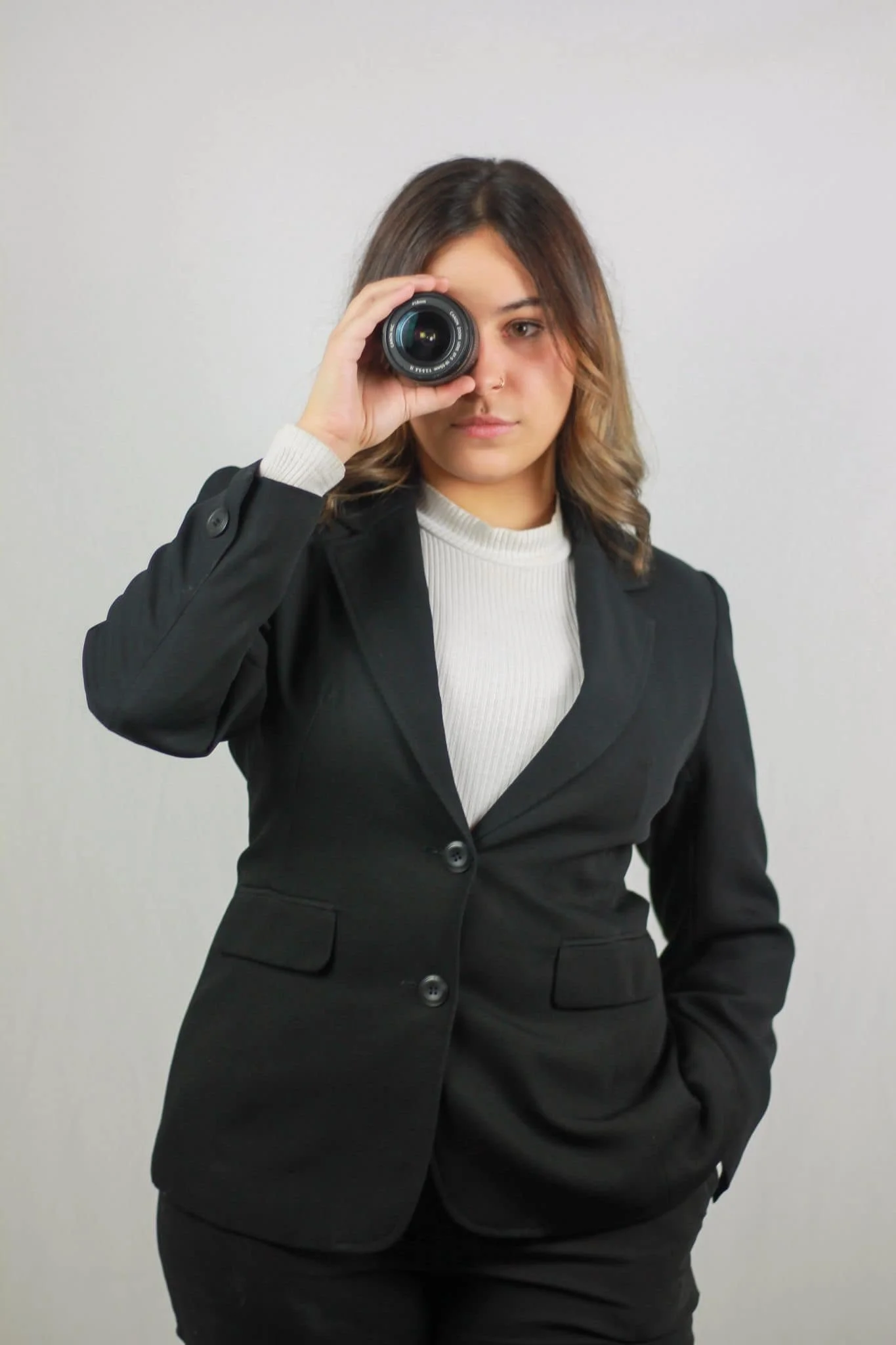 A woman with shoulder-length wavy brown hair holding a camera lens up to her right eye, dressed in a black blazer over a white ribbed turtleneck, standing against a plain white background.