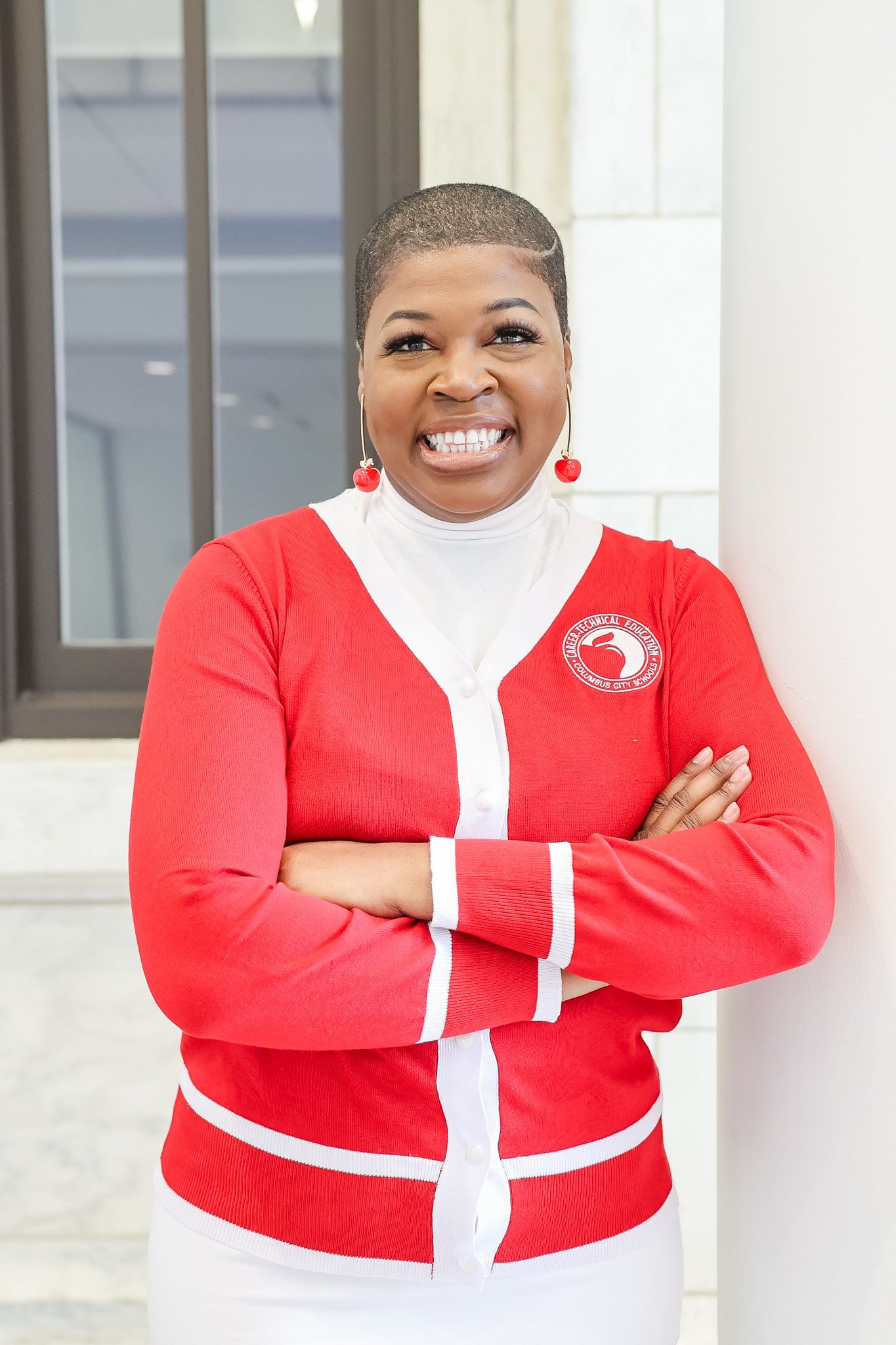 A woman with a short hairstyle smiling in a bright indoor setting, wearing a red cardigan with white detailing and a school emblem, and cherry-shaped earrings.