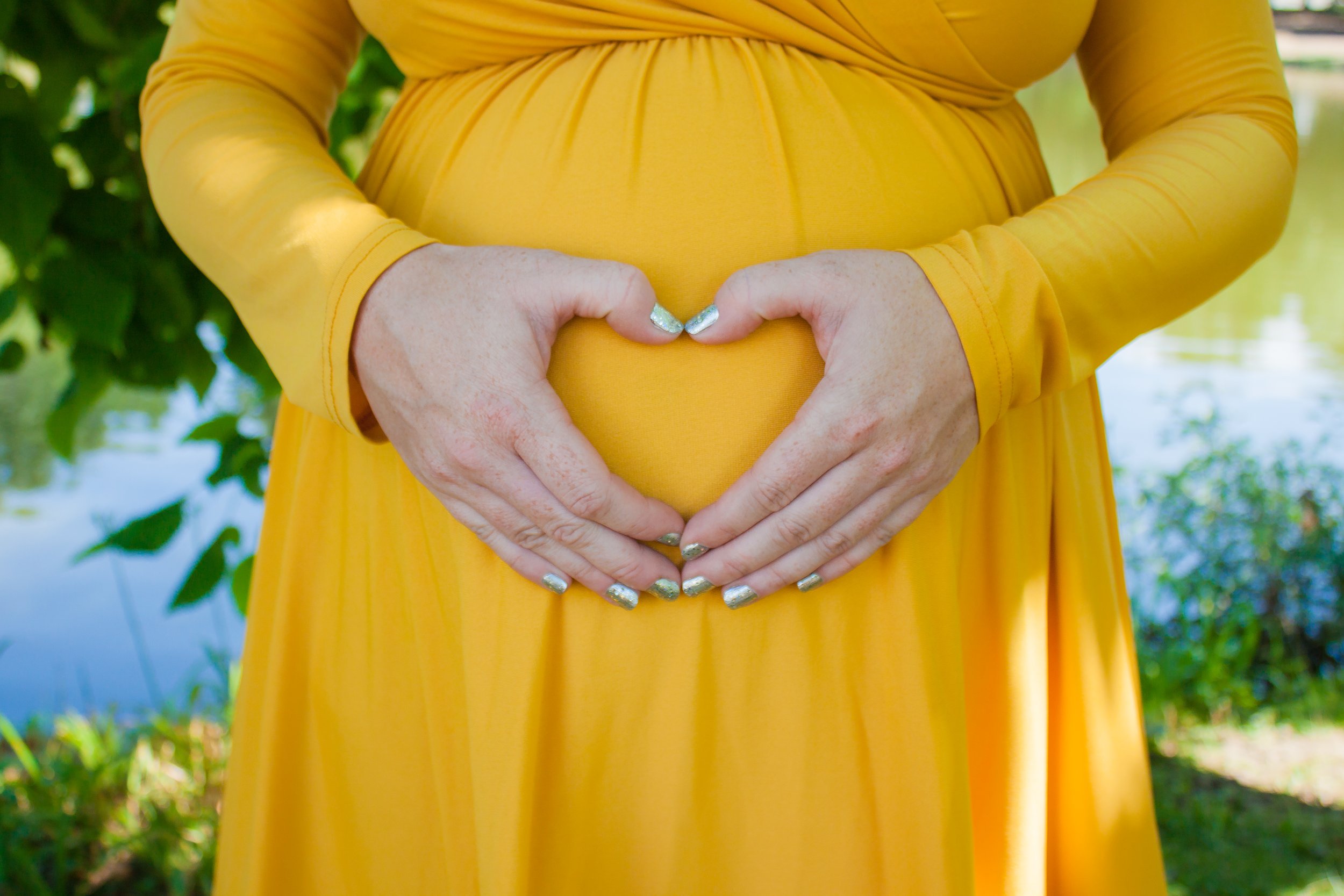 Person wearing a yellow dress making a heart shape with hands over a pregnant belly.