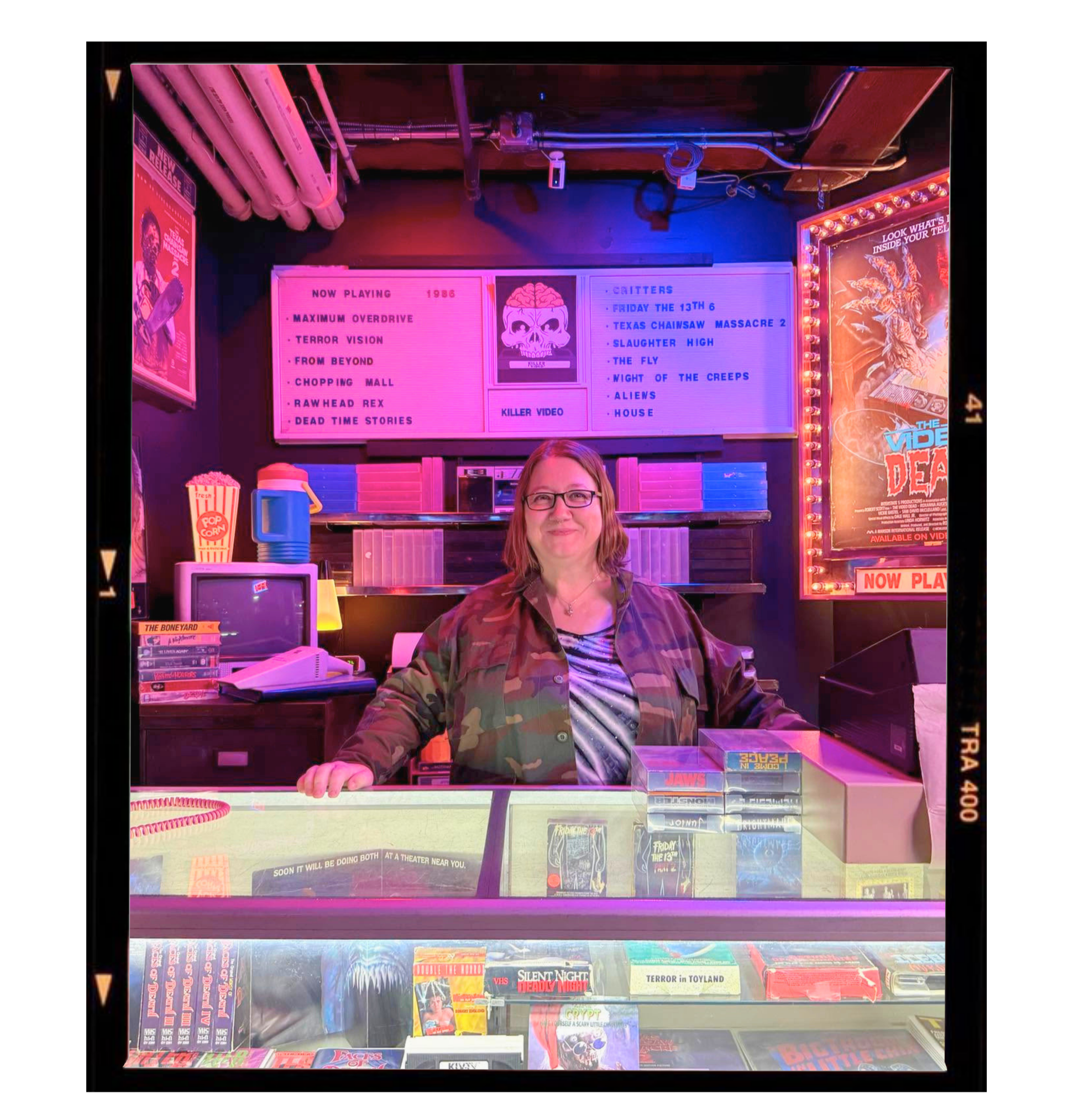 A woman with glasses and brown hair standing behind a glass counter of DVDs and VHS tapes in a store, with neon pink and purple lighting, and movie posters on the walls.
