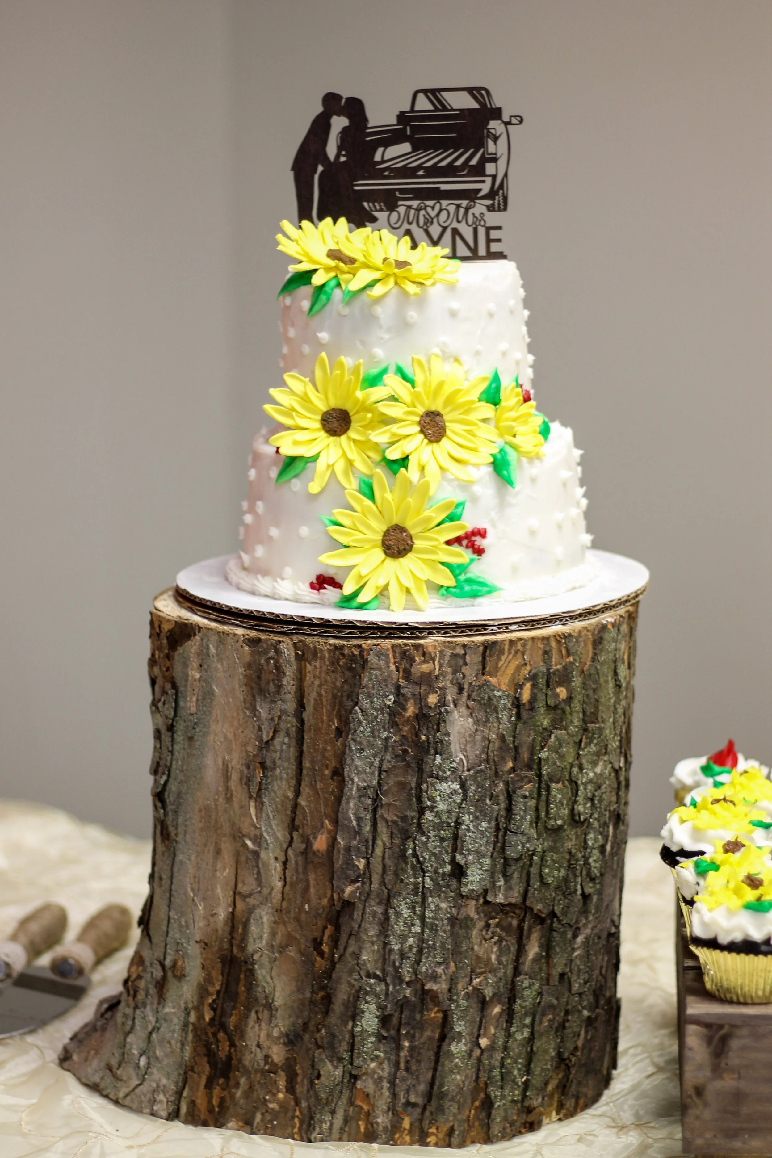 A wedding cake decorated with yellow sunflowers and a topper featuring a couple kissing near a piano, placed on a large tree stump.