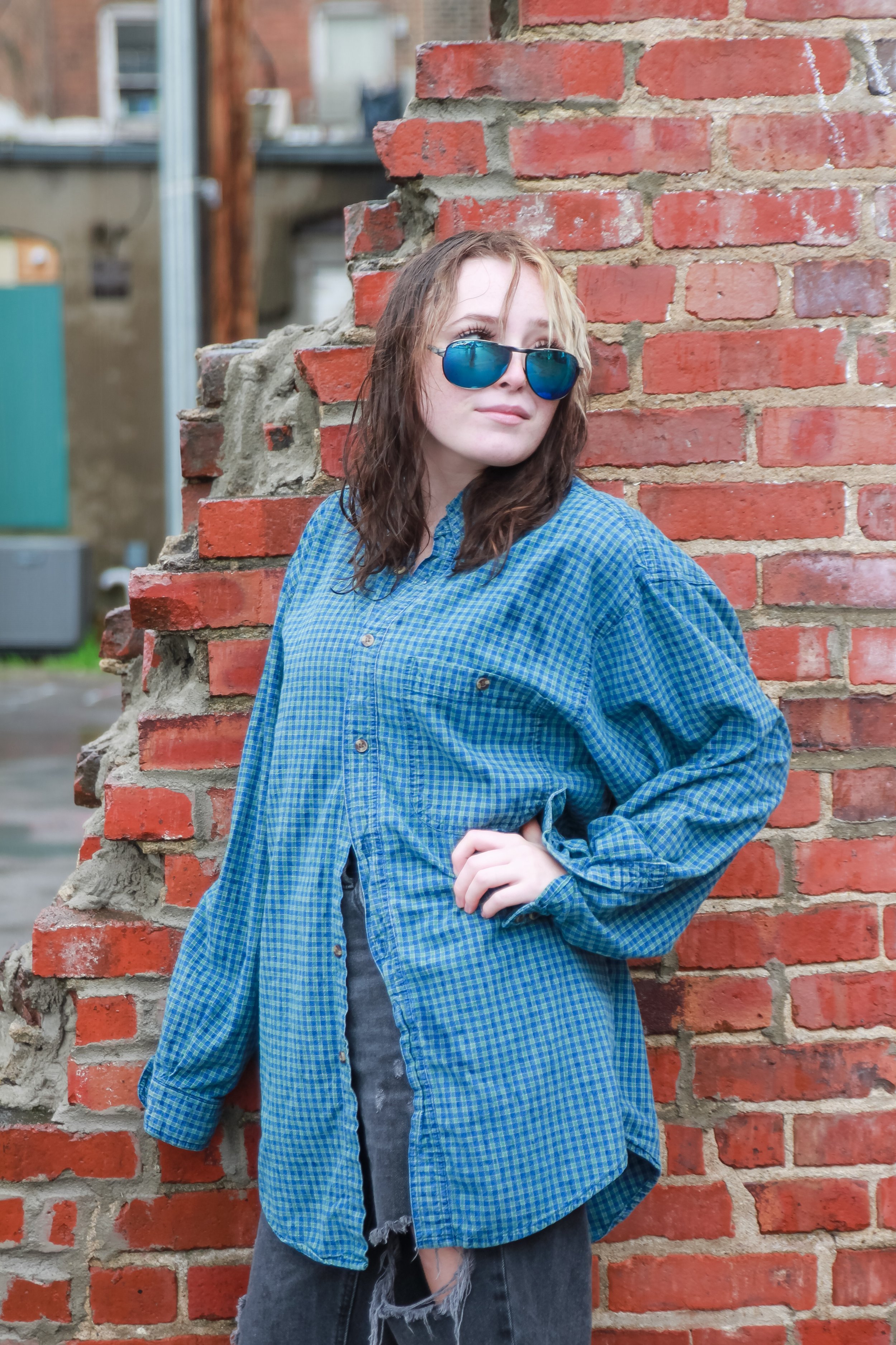 A young woman with wavy brown hair wearing dark sunglasses, a blue checkered shirt, and black ripped jeans, leaning against a partially ruined brick wall in an urban outdoor setting.