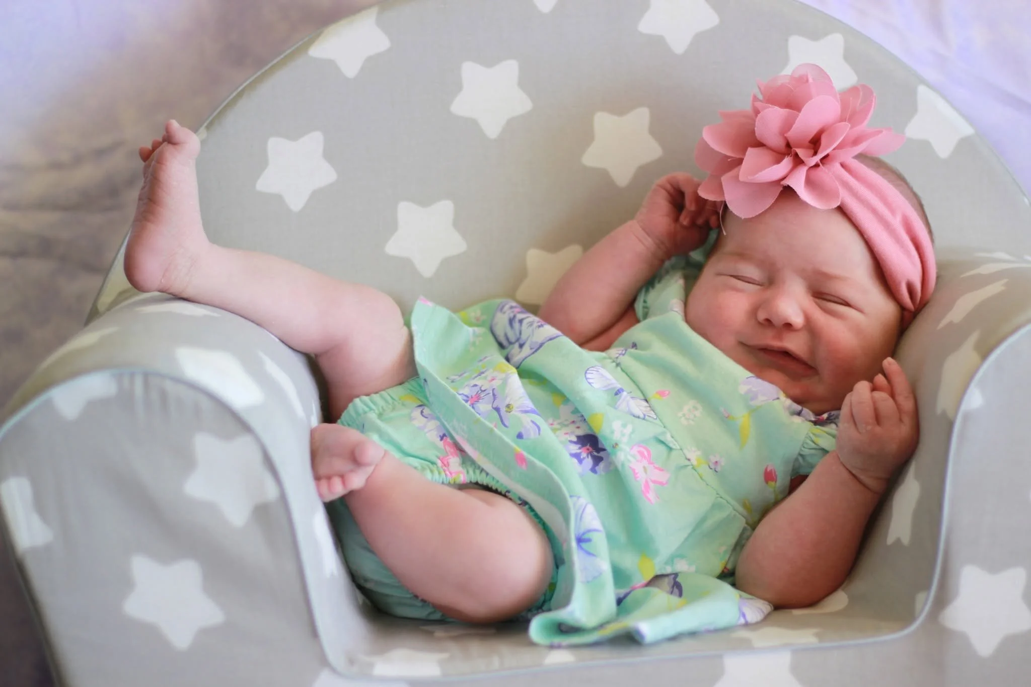 A newborn baby sleeping in a bassinet, wearing a pink hat and a colorful onesie with a floral pattern, inside a star-patterned bassinet.