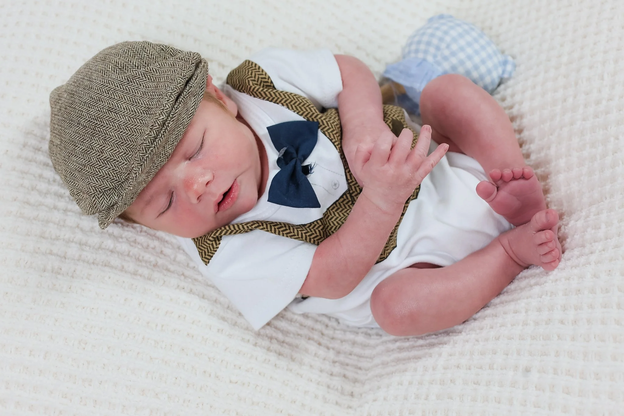 A realistic baby doll lying on a white textured surface, wearing a brown and black herringbone cap, a white outfit with a blue bow, and blue checkered socks.