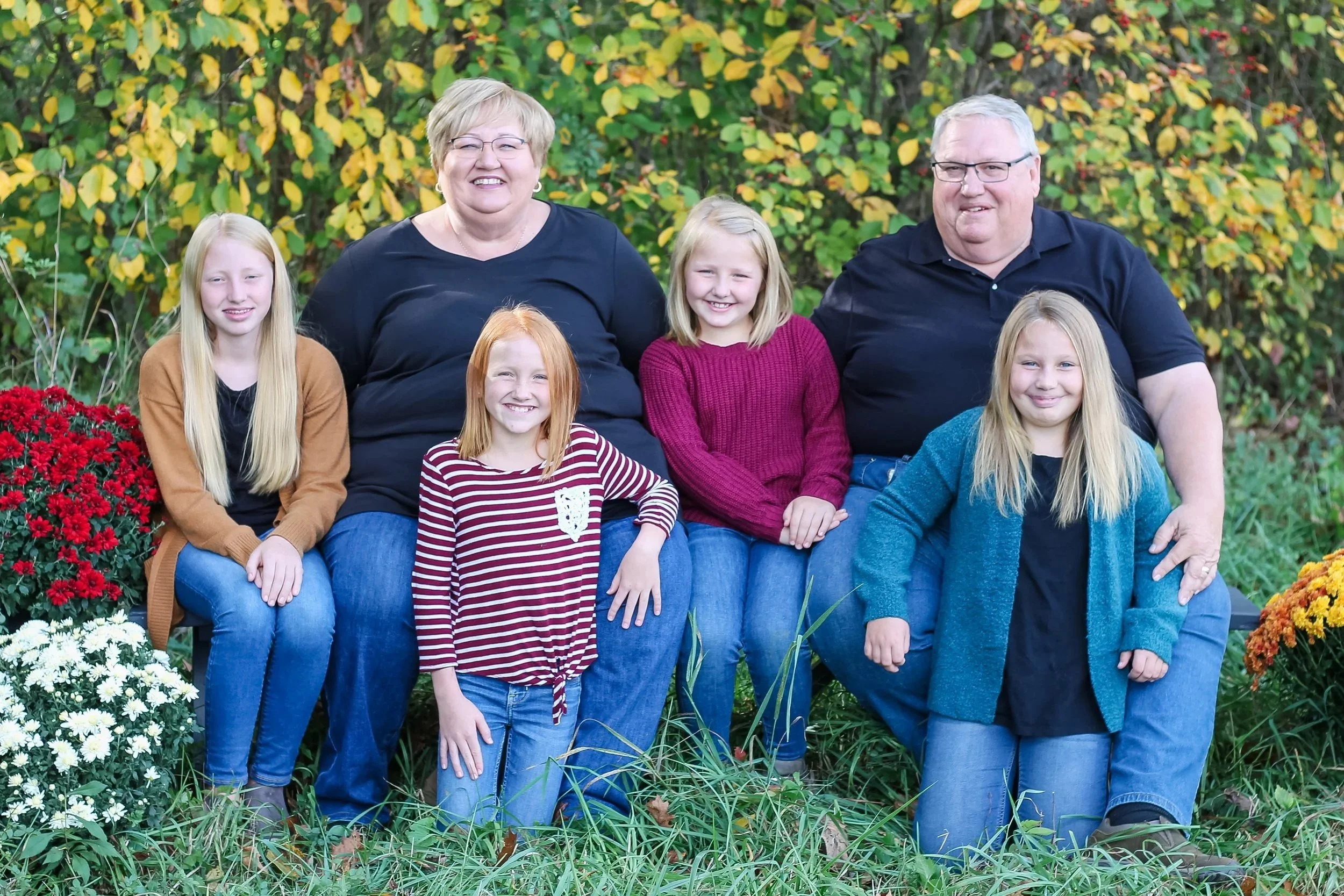 A family of seven, including grandparents, parents, and children, sitting together outdoors on a bench surrounded by fall foliage and colorful flowers, smiling at the camera.