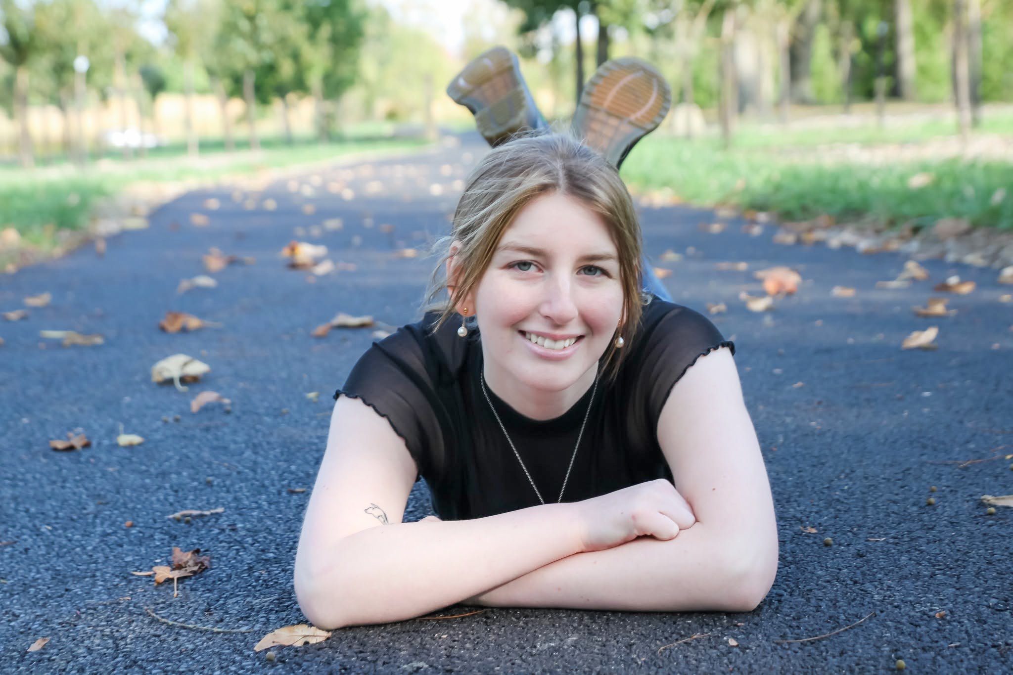 A young woman with light brown hair lying on her stomach on a paved path in a park, smiling at the camera. She is wearing a black sleeveless top, earrings, and a necklace, with her arms crossed in front of her. The background shows trees and fallen l