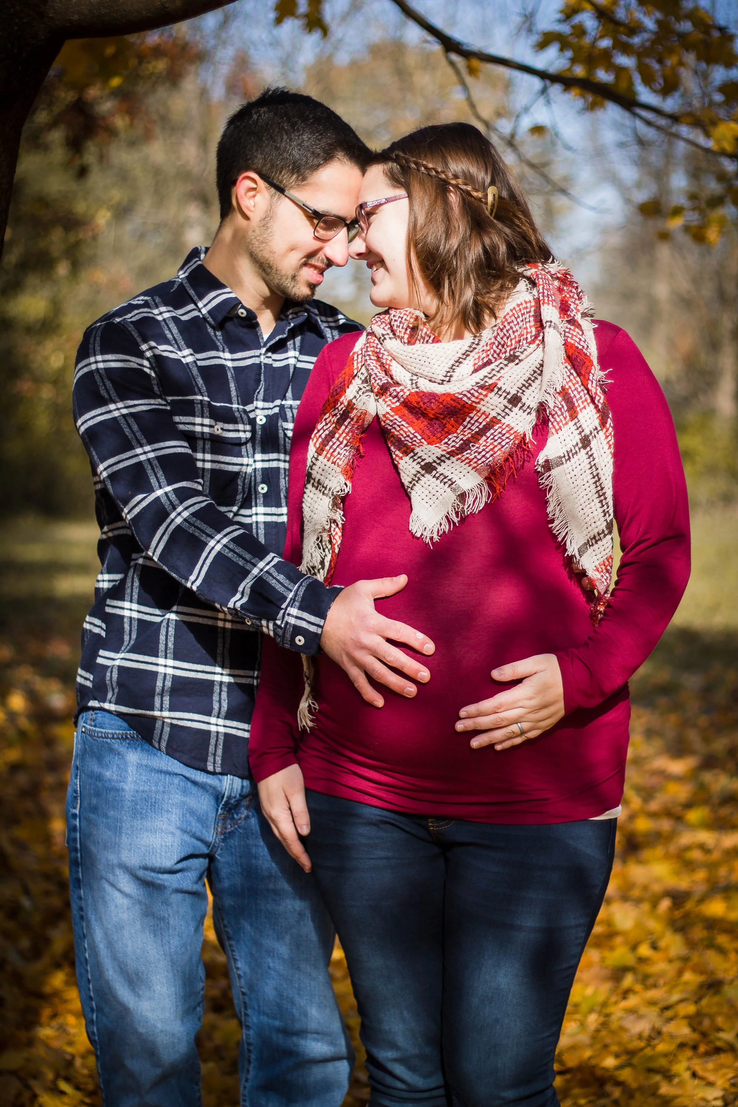 A couple standing close together outdoors in autumn, with their foreheads touching and hands on the pregnant woman's belly, smiling.