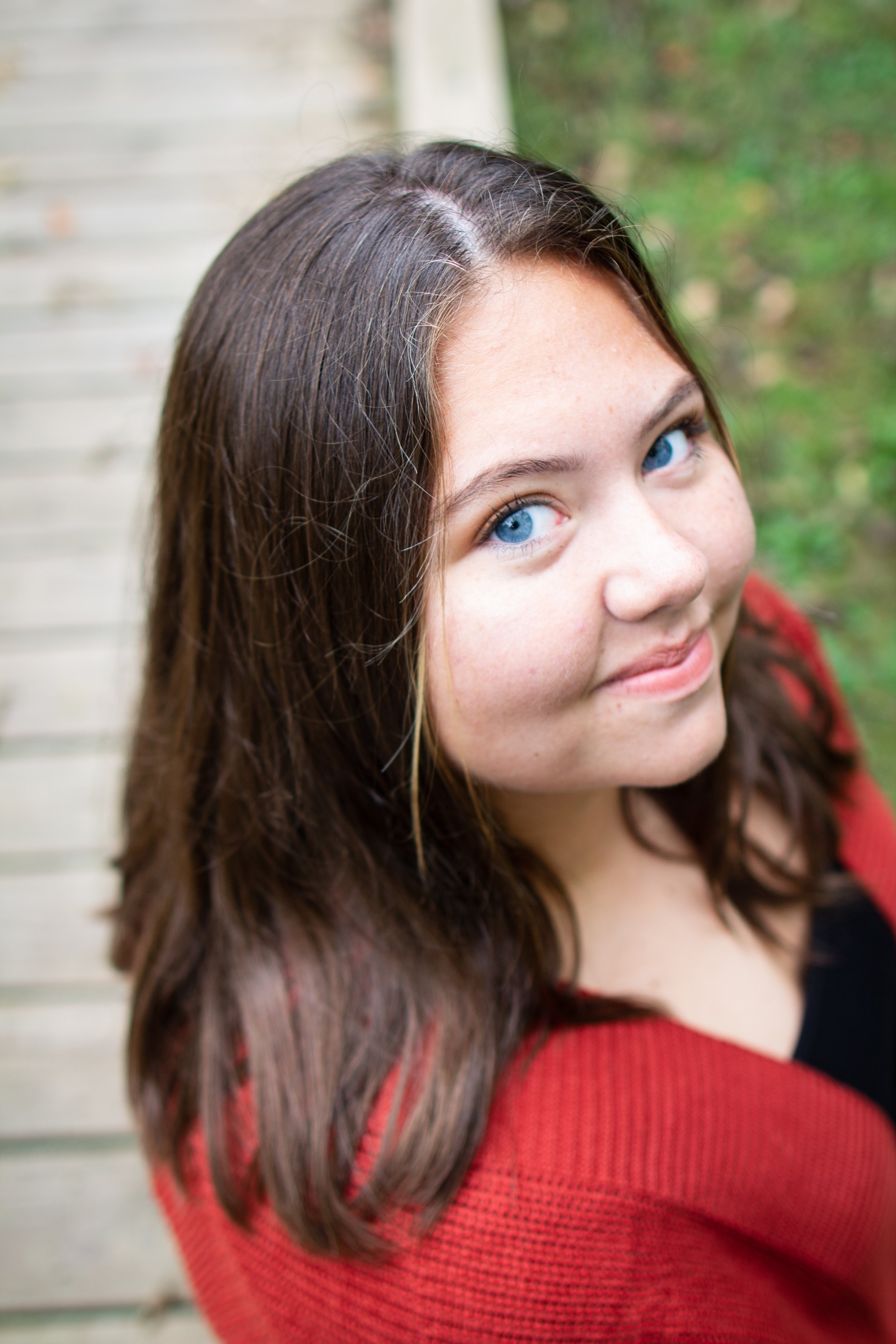 Close-up of a woman with blue eyes and long brown hair wearing a red and black sweater, smiling outdoors with greenery in the background.