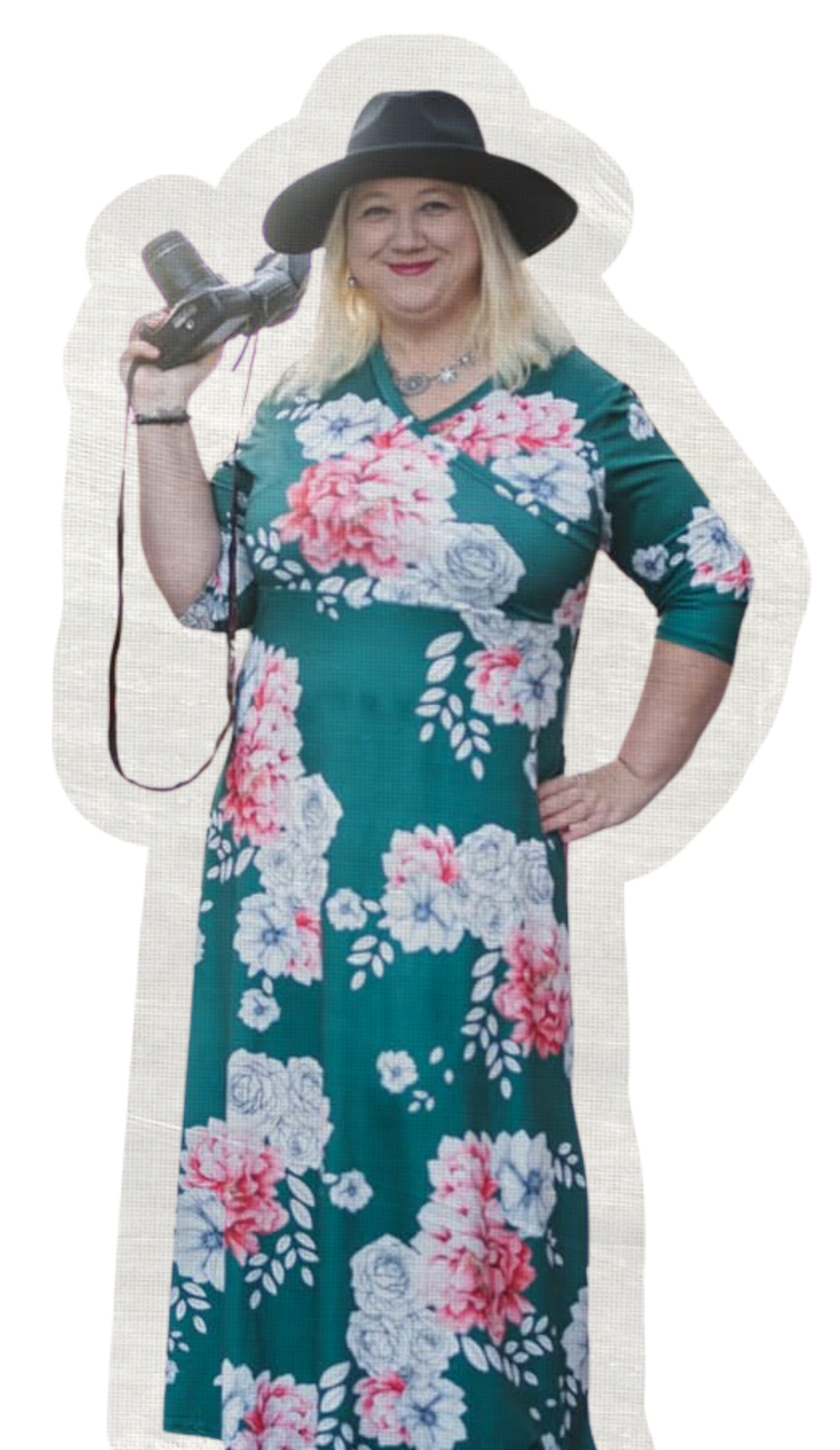 A woman with blonde hair wearing a black wide-brimmed hat and green floral dress holding a camera, smiling at the camera.