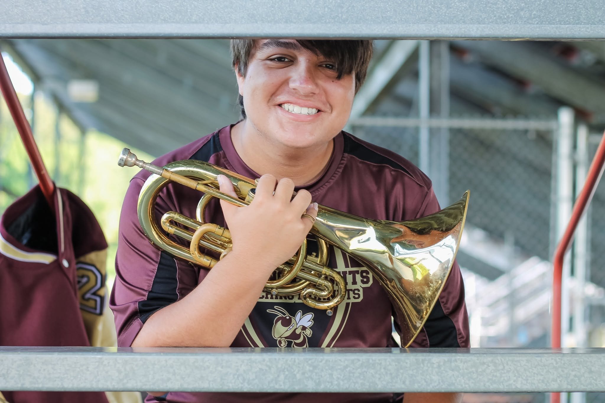 A young man with brown hair smiling while holding a brass trumpet, wearing a maroon sports jersey, standing behind a metal railing at a sports event.
