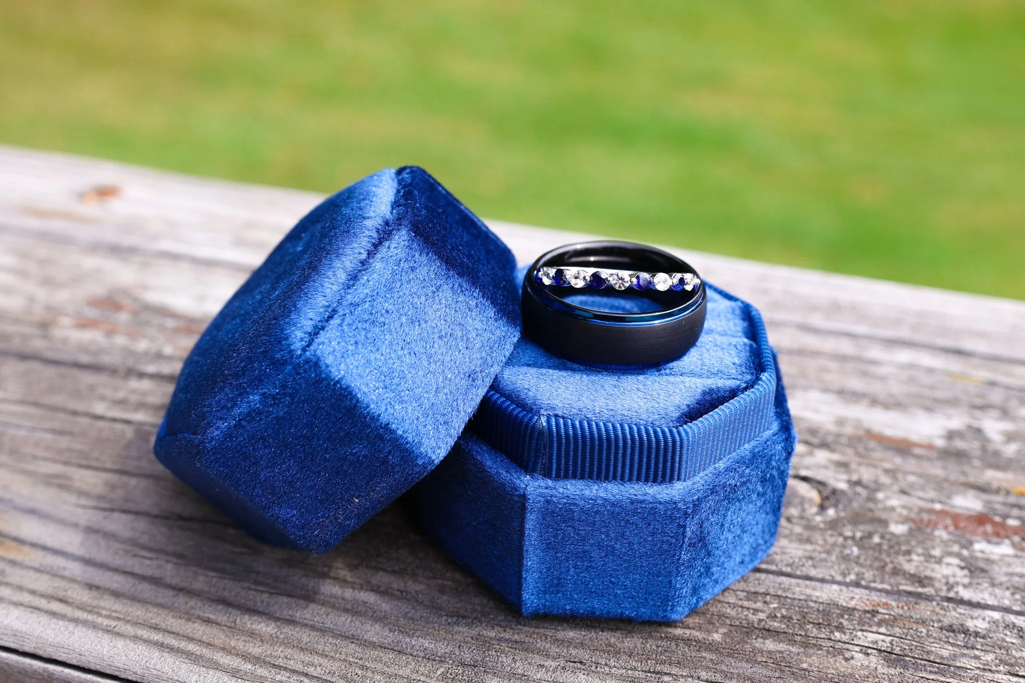 A black ring with a row of small clear gemstones, placed inside a blue velvet ring box resting on a blue velvet pouch on a wooden surface.