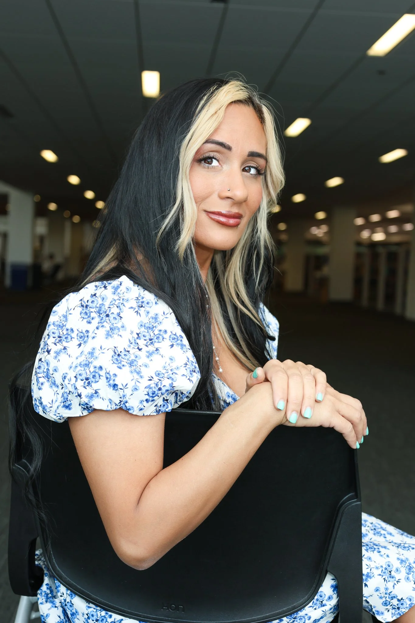 A woman with long black and blonde hair, wearing a blue and white floral dress, is sitting at a black table, resting her hands together, and looking at the camera with a slight smile. The background shows an indoor setting with ceiling lights.