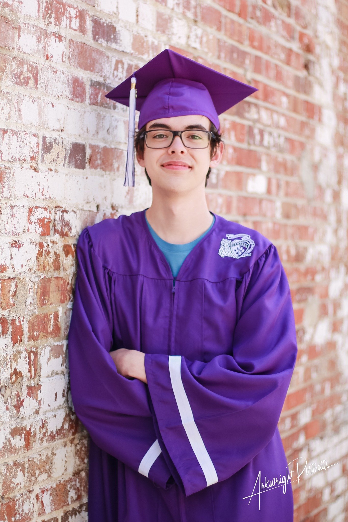 Young man in purple graduation gown and cap with a tassel, standing with arms crossed in front of a brick wall.
