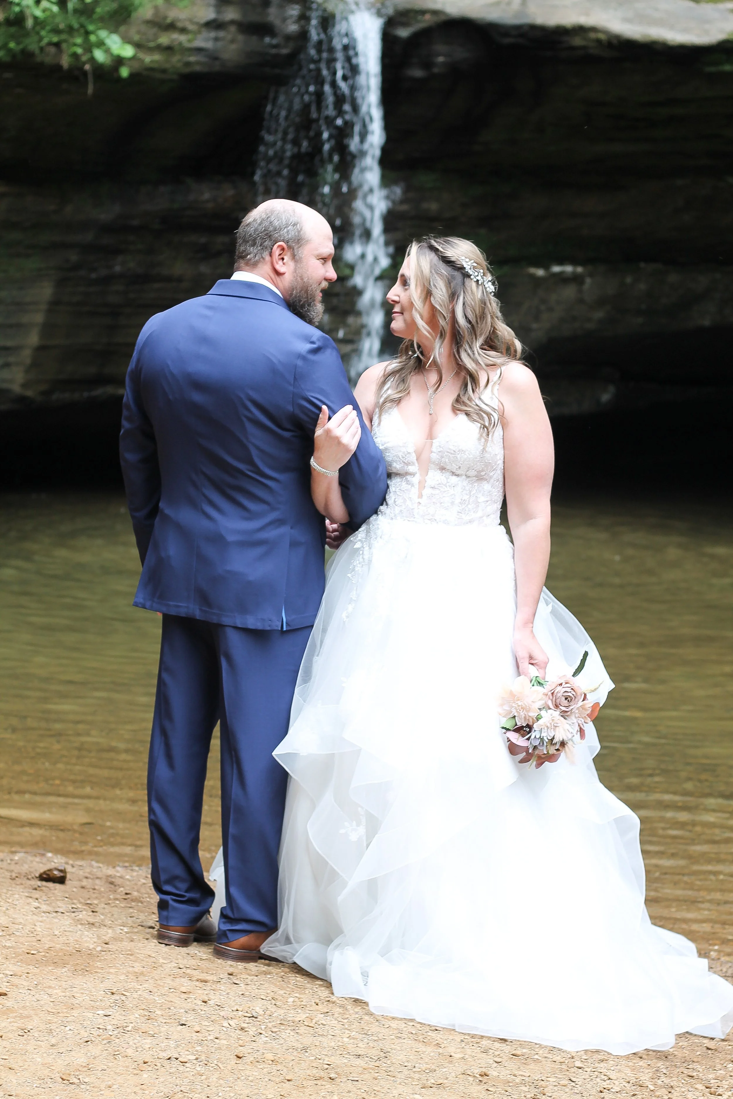 A bride and groom standing close together outdoors near a waterfall, with the bride holding a bouquet of flowers.