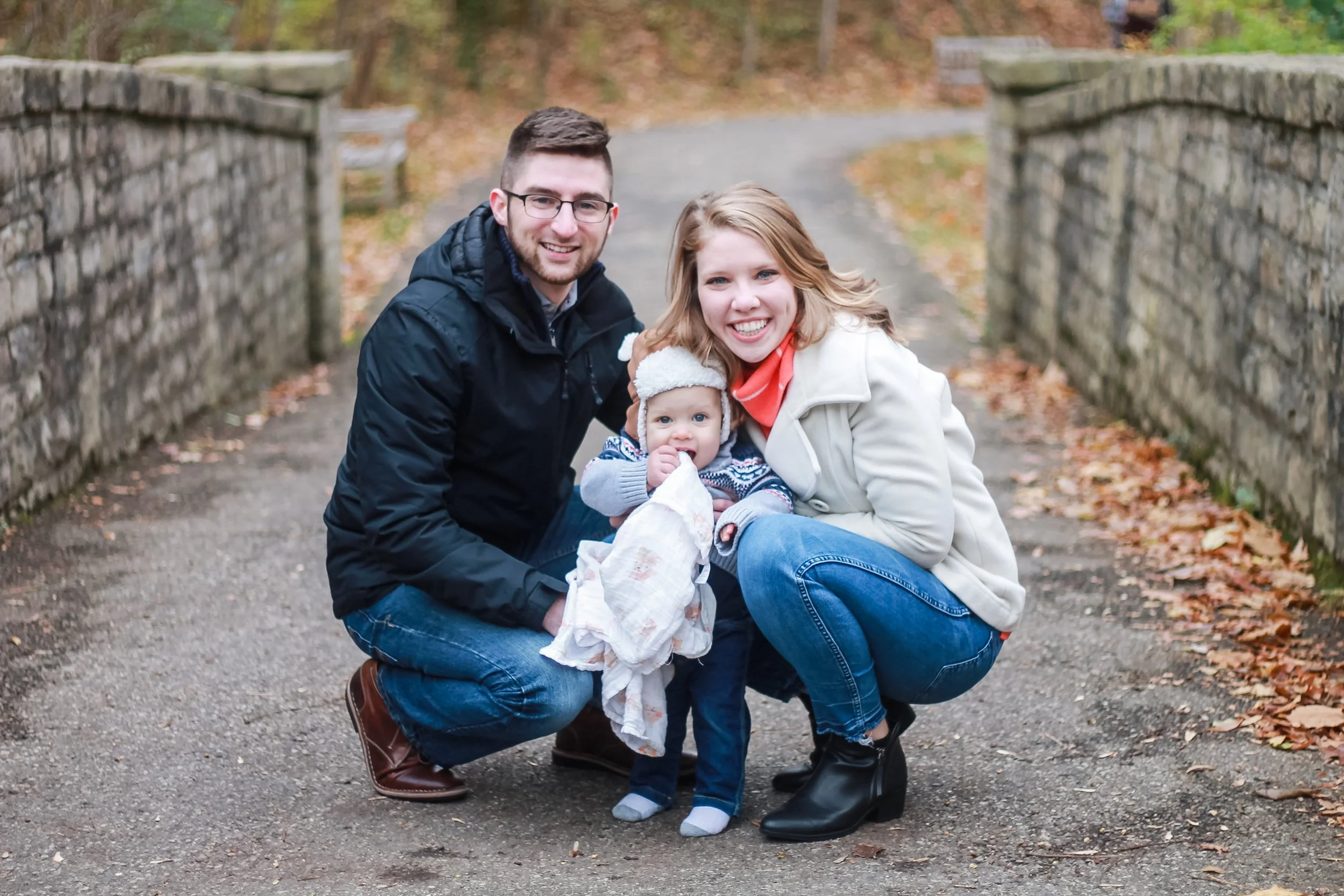 A happy family of three, a man, a woman, and a young girl, crouching on a paved path in an outdoor park during fall, smiling at the camera with autumn leaves scattered on the ground.