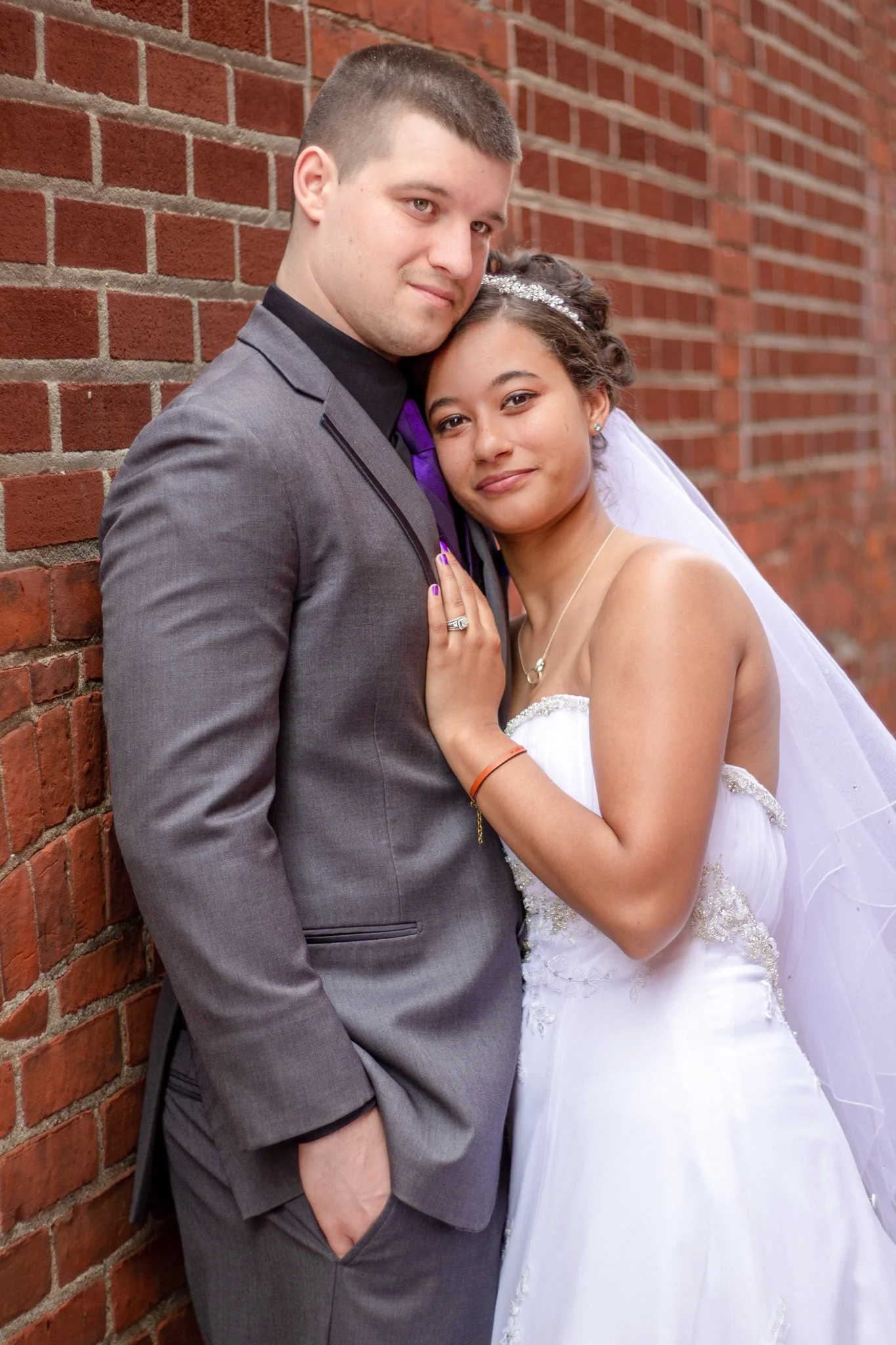 A newlywed couple leaning against a brick wall, the groom wearing a gray suit and the bride in a white wedding dress with a tiara and veil, looking at the camera.