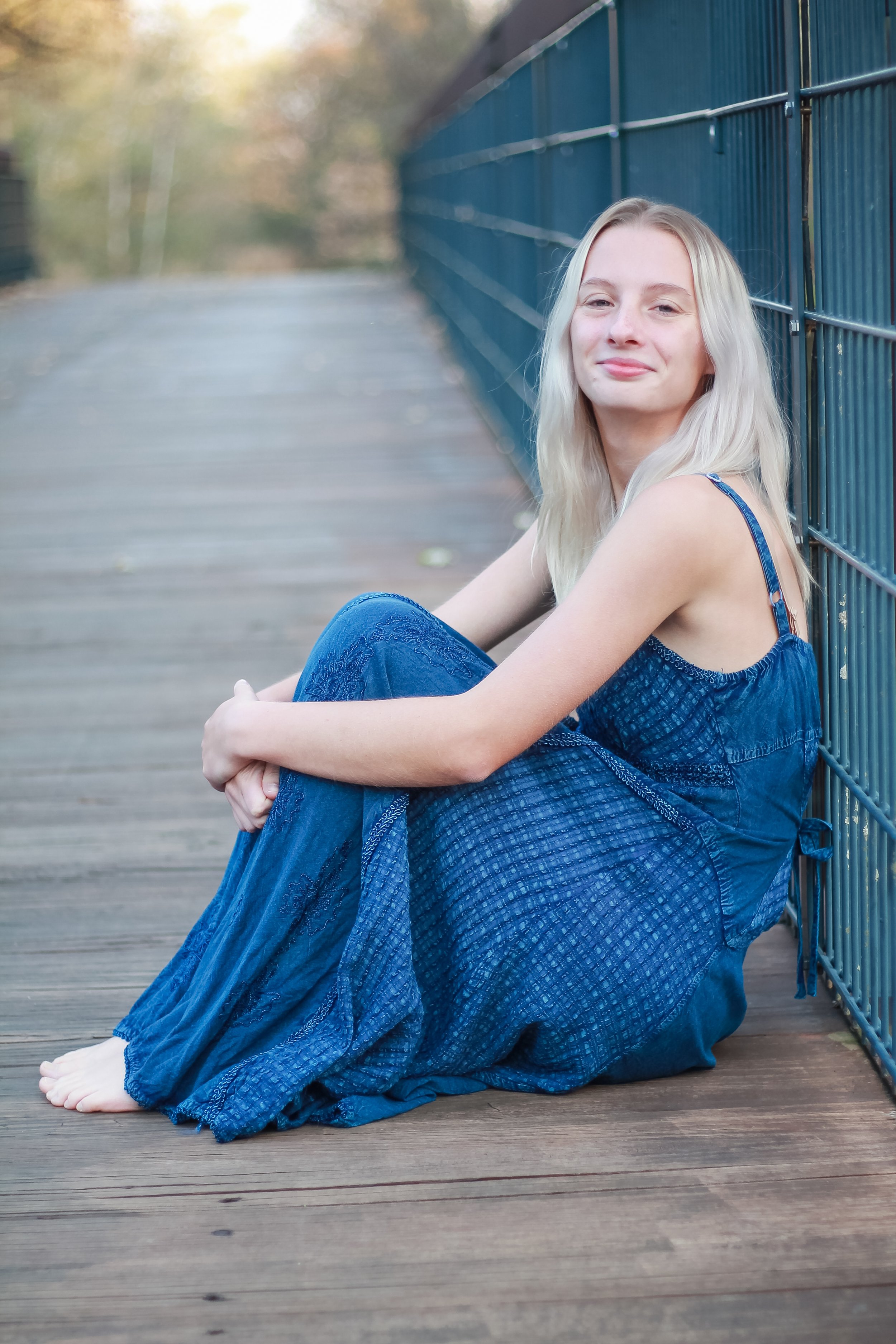 A young woman with long blonde hair sitting on a wooden deck, leaning against a blue metal fence with a blurred background of trees and sky.