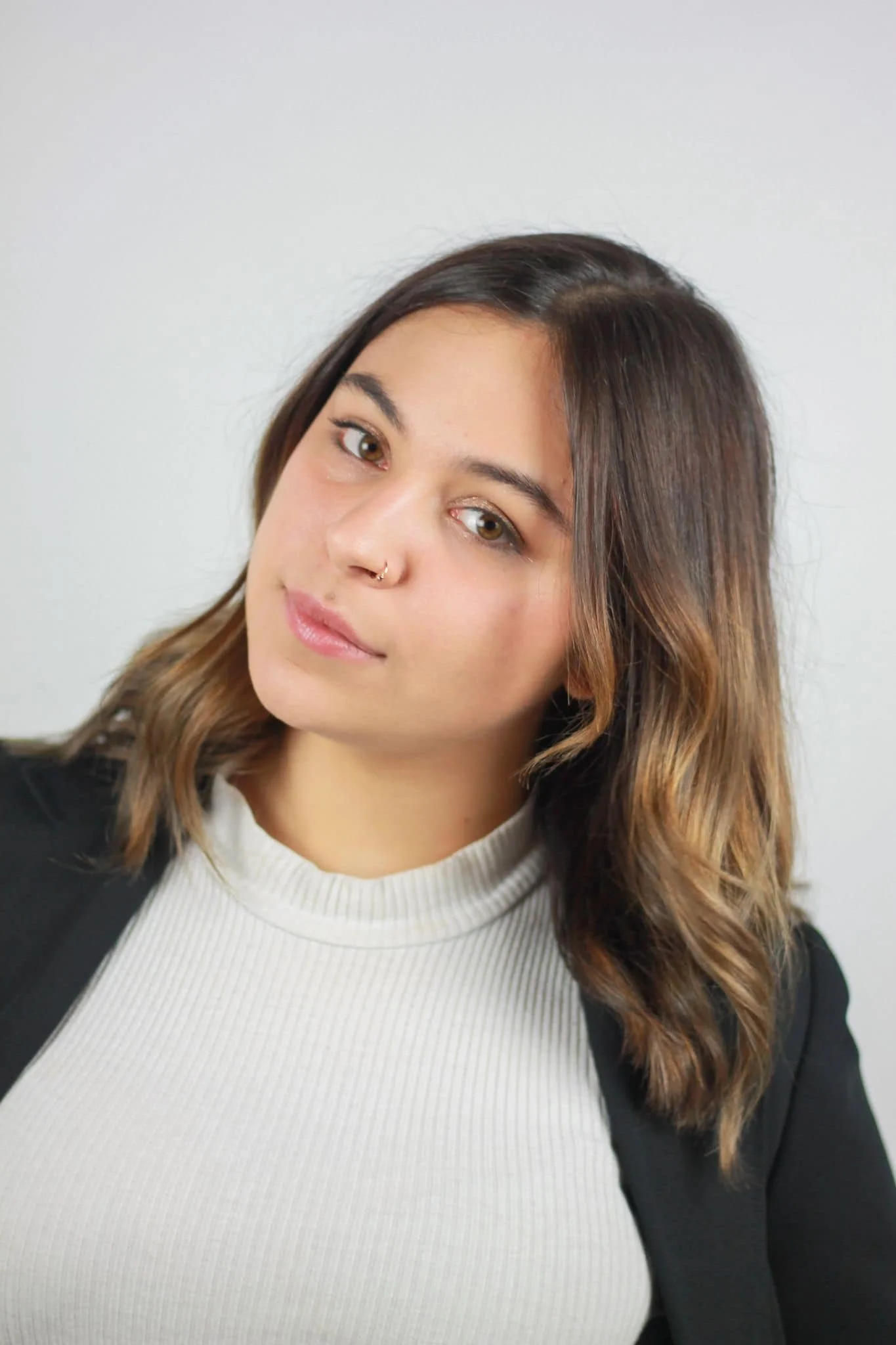 Close-up of a young woman with wavy brown hair, wearing a white ribbed turtleneck sweater and a black blazer, looking at the camera with a slight smile.