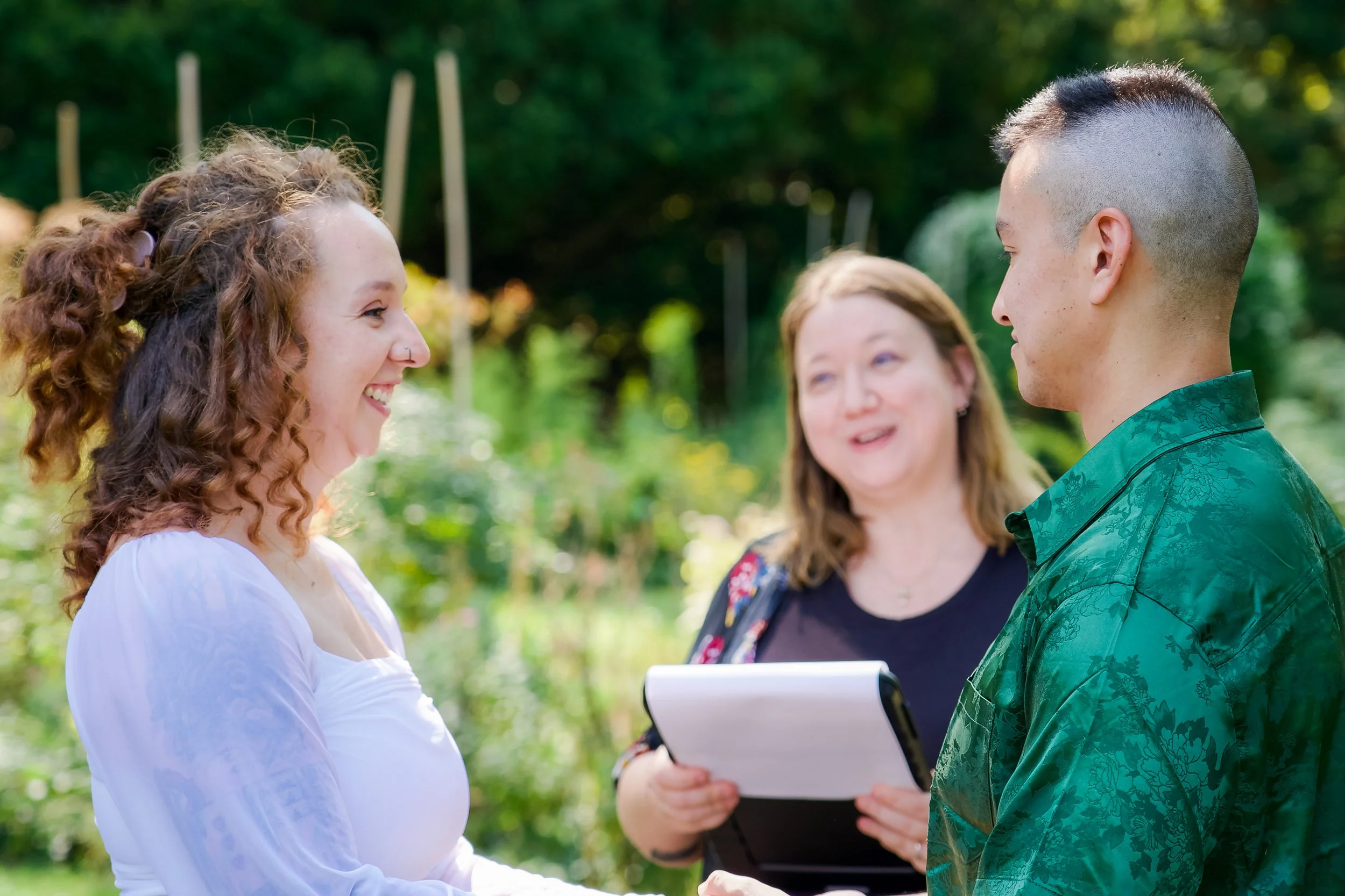 A woman with curly red hair and a nose ring smiling at a man with a mohawk hairstyle during a wedding ceremony outdoors, with an officiant holding a book in the background.