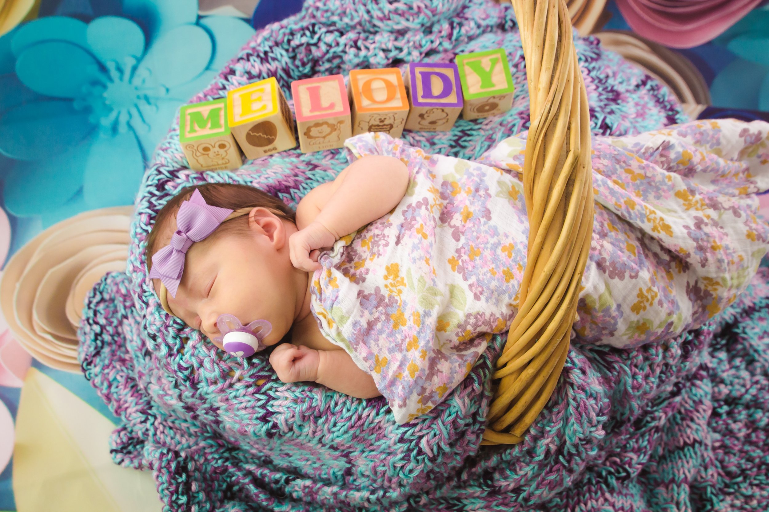 A sleeping baby lying in a wicker basket, wearing a floral dress and a purple bow headband. The basket is lined with a multicolored knitted blanket. Colorful blocks spell out 'MELODY' behind the baby, with images of a bear, a lion, and a monkey on so