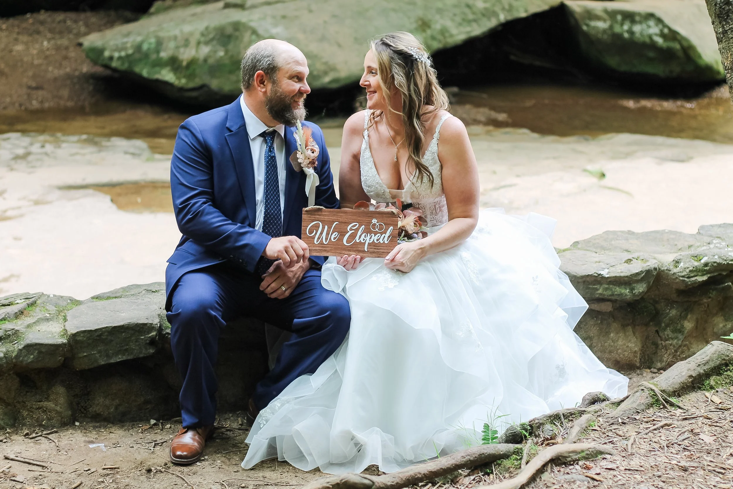 A bride and groom sitting on rocks outdoors near a stream, holding a wooden sign that says 'We Eloped' and smiling at each other.