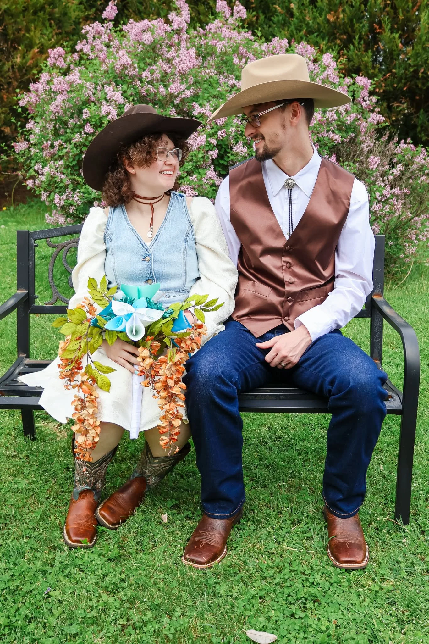 A man and woman dressed as cowboys sitting on a park bench, smiling and looking at each other. The woman is holding a bouquet of colorful paper flowers and wearing cowboy boots, jeans, a white blouse, and a cowboy hat. The man is wearing a white shirt, a brown vest, blue jeans, boots, and a cowboy hat. There is a large bush with pink flowers behind them and grass on the ground.