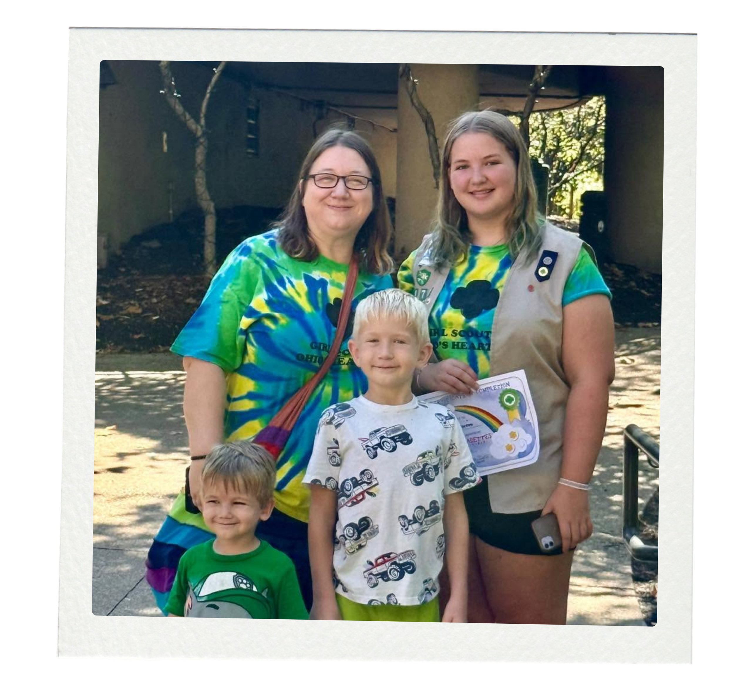 Four people, including two children, standing outdoors. The woman on the left is wearing glasses and a colorful tie-dye shirt. The young girl on the right is wearing a scout uniform with badges. The two boys are in front, one wearing a patterned shirt, and the other a green shirt with a rainbow design. All are smiling.