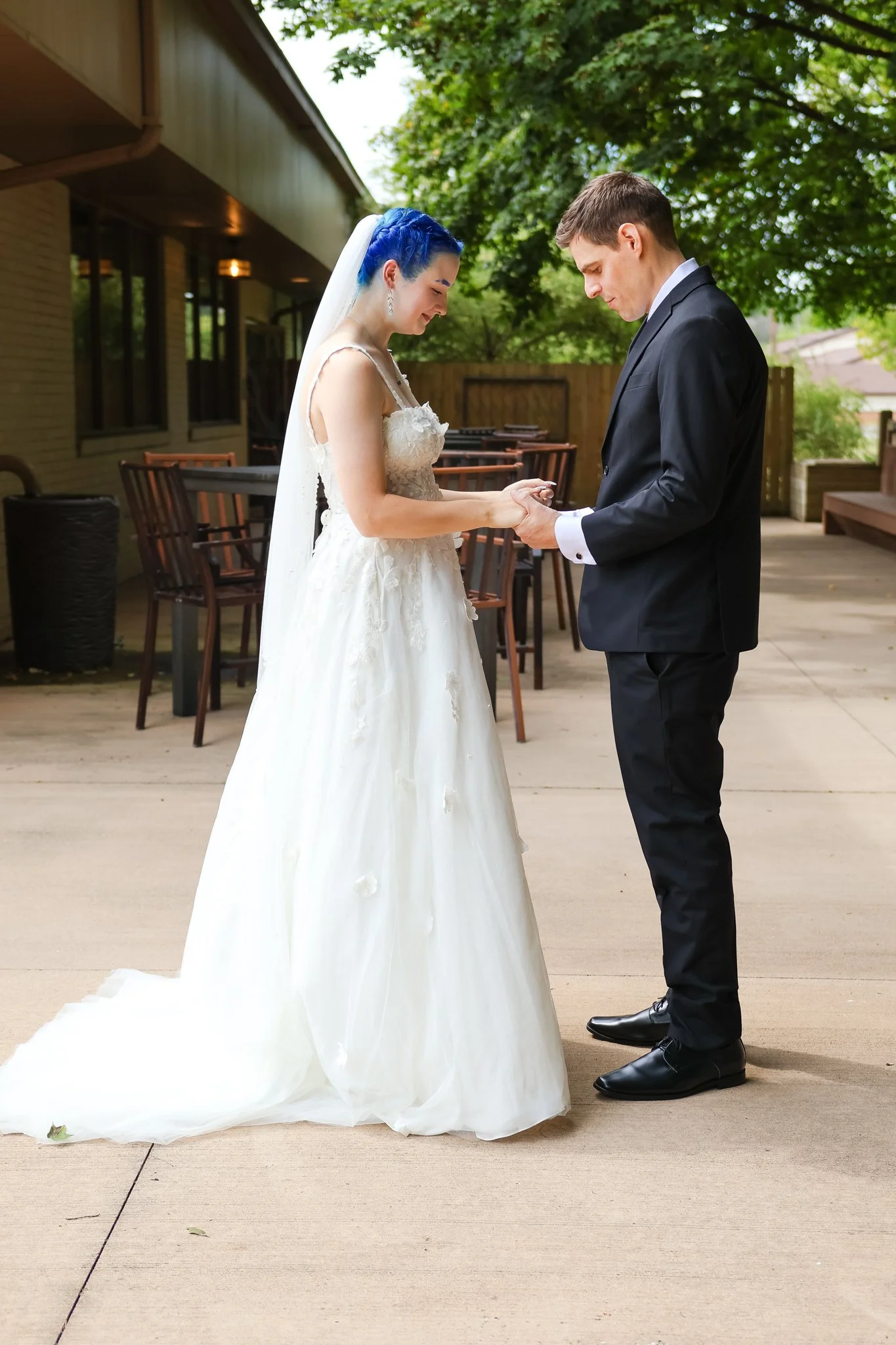 A bride and groom stand outside during the daytime, holding hands and looking at each other. The bride has blue hair and is wearing a white wedding dress with a long train, and the groom is dressed in a black tuxedo. They are under a canopy of green 