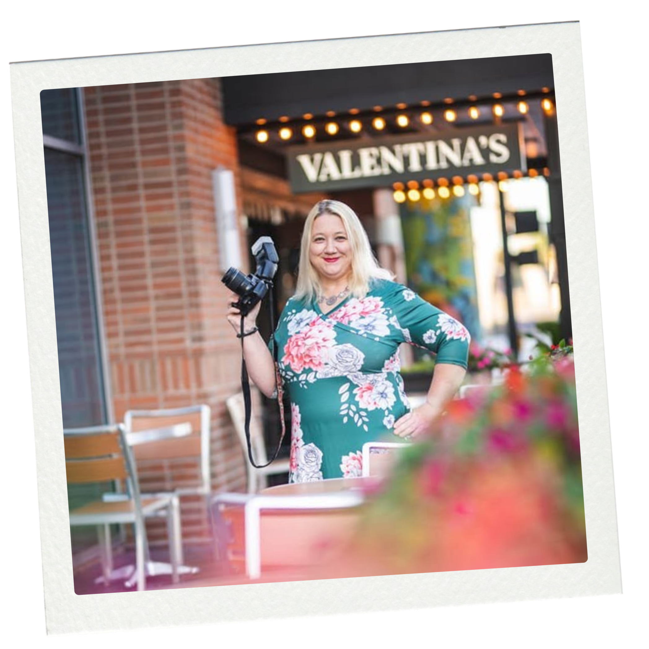 A woman with blonde hair holding a camera, standing outside near a brick building with an illuminated sign that says 'Valentina's' and string lights overhead.