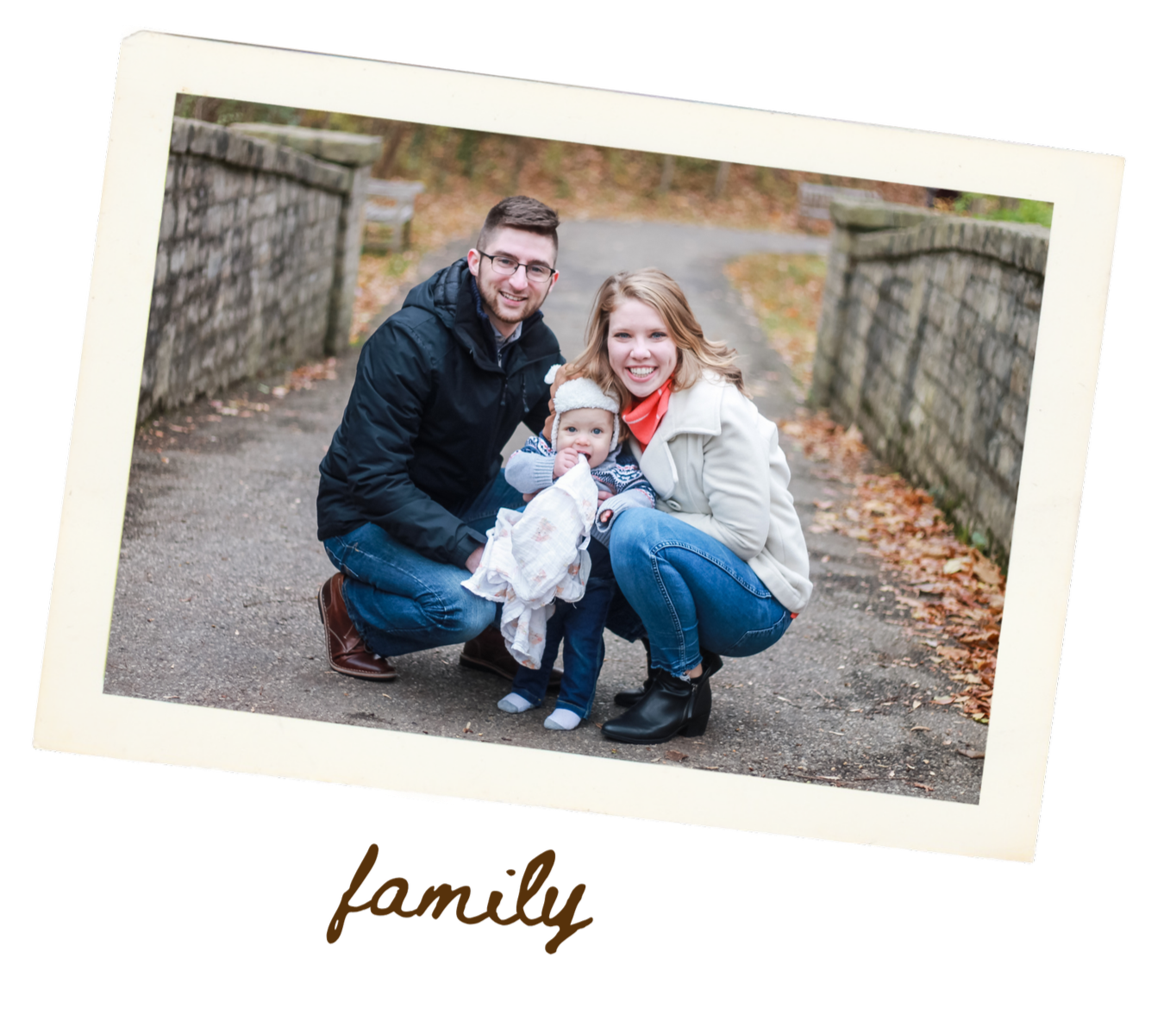 A family of three posing outdoors on a path with stone walls, autumn leaves on the ground, smiling at the camera. The father and mother are crouching on each side of their young child, who is holding a blanket and wearing a knit hat.