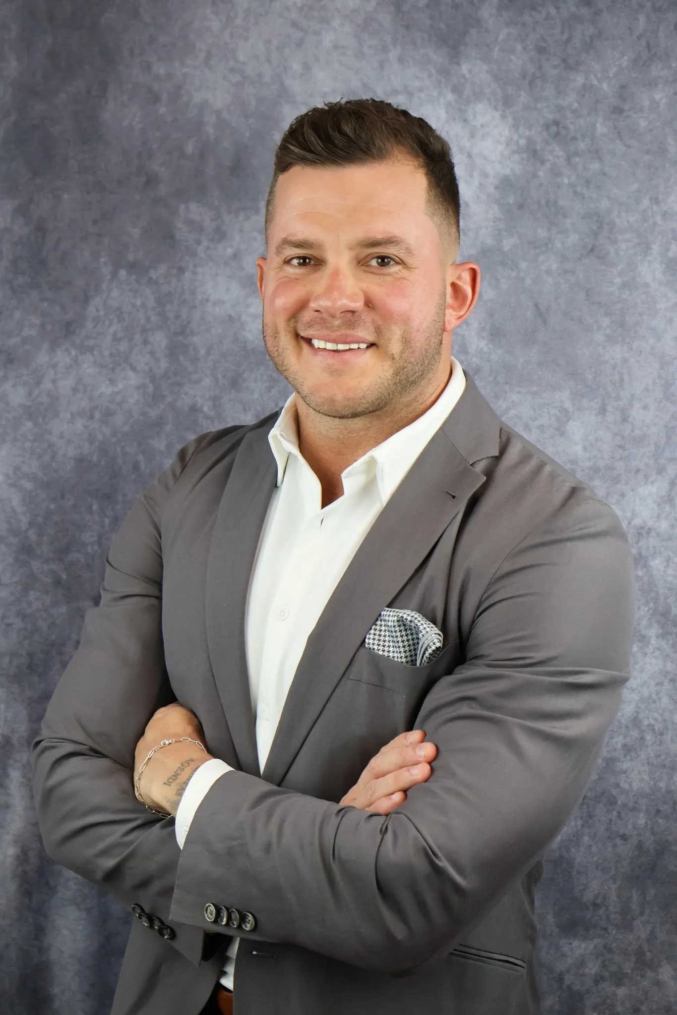 Smiling man in a gray suit with gray and white patterned pocket square, white shirt, and silver bracelet, standing against a textured gray background.