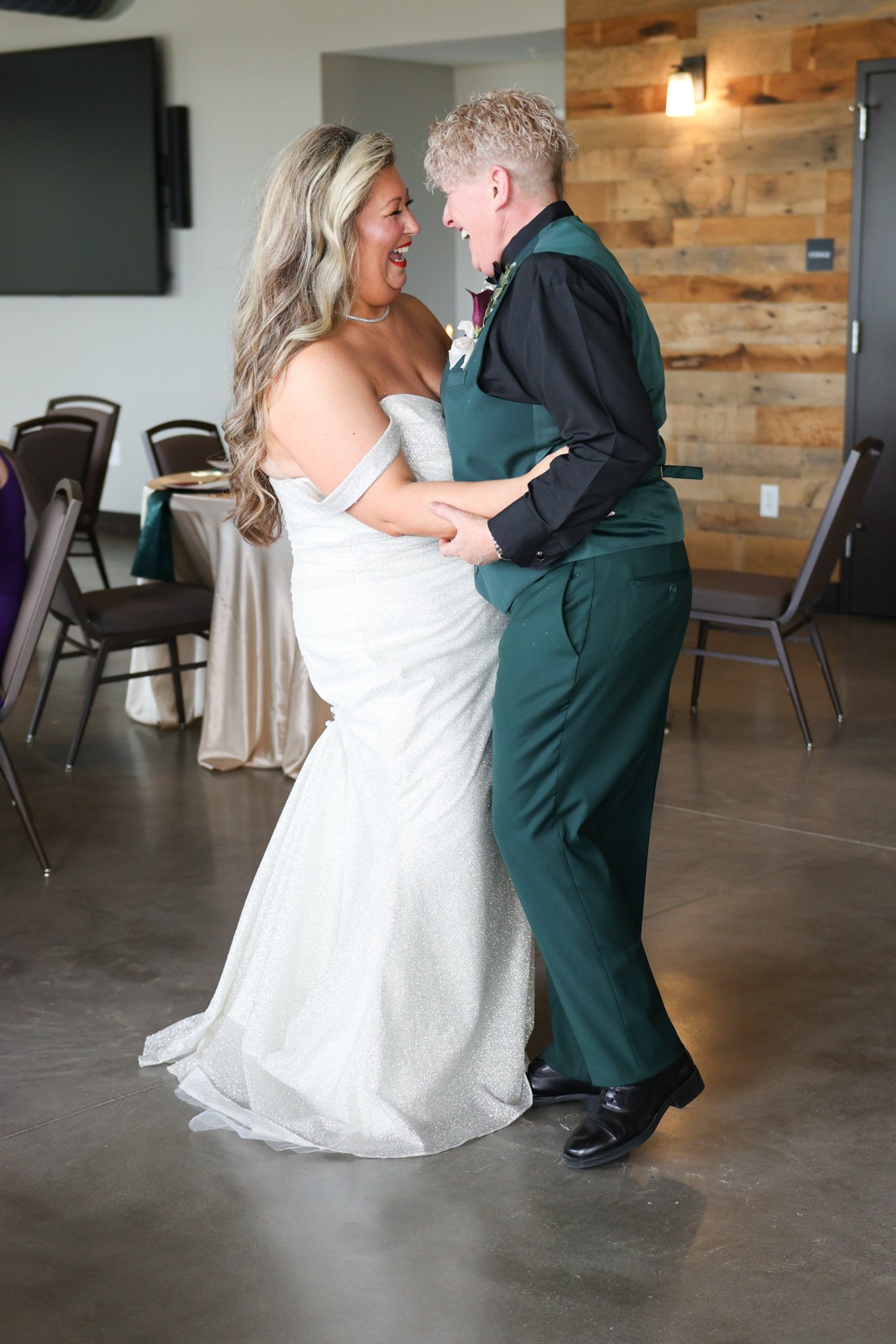 A couple dancing at a wedding reception. The bride is wearing a white wedding gown, and the groom is dressed in a dark suit with green accents. They are smiling and looking at each other, holding hands while dancing. The background includes chairs and a wooden accent wall.