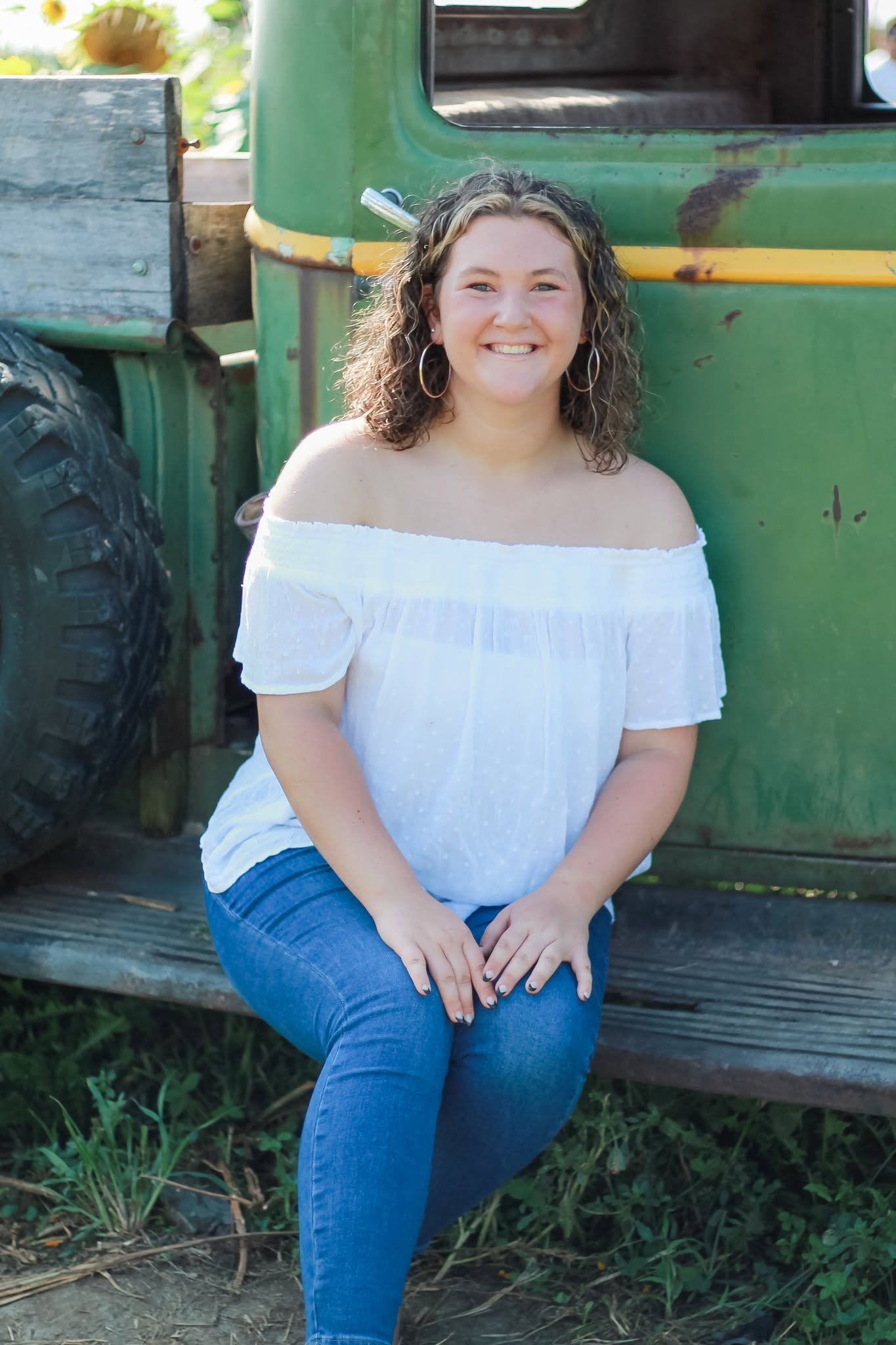 A woman with curly hair wearing a white off-the-shoulder top and blue jeans, sitting on a wooden bench in front of a green vintage truck.