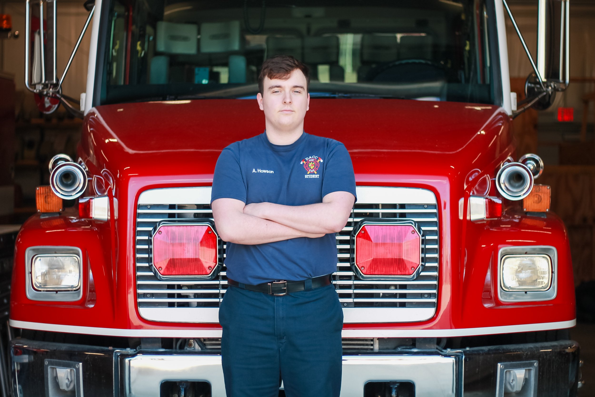 A young man with short brown hair standing with arms crossed in front of a red fire truck inside a garage. He is wearing a navy blue t-shirt with an emblem and the words 'A. Howson' and 'STUDENT' on it.
