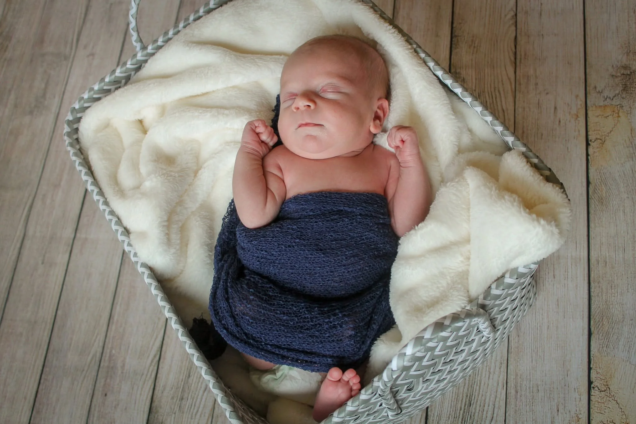 A sleeping newborn baby wrapped in a dark blue blanket, lying in a white basket lined with soft, cream-colored fabric, on a wooden floor.