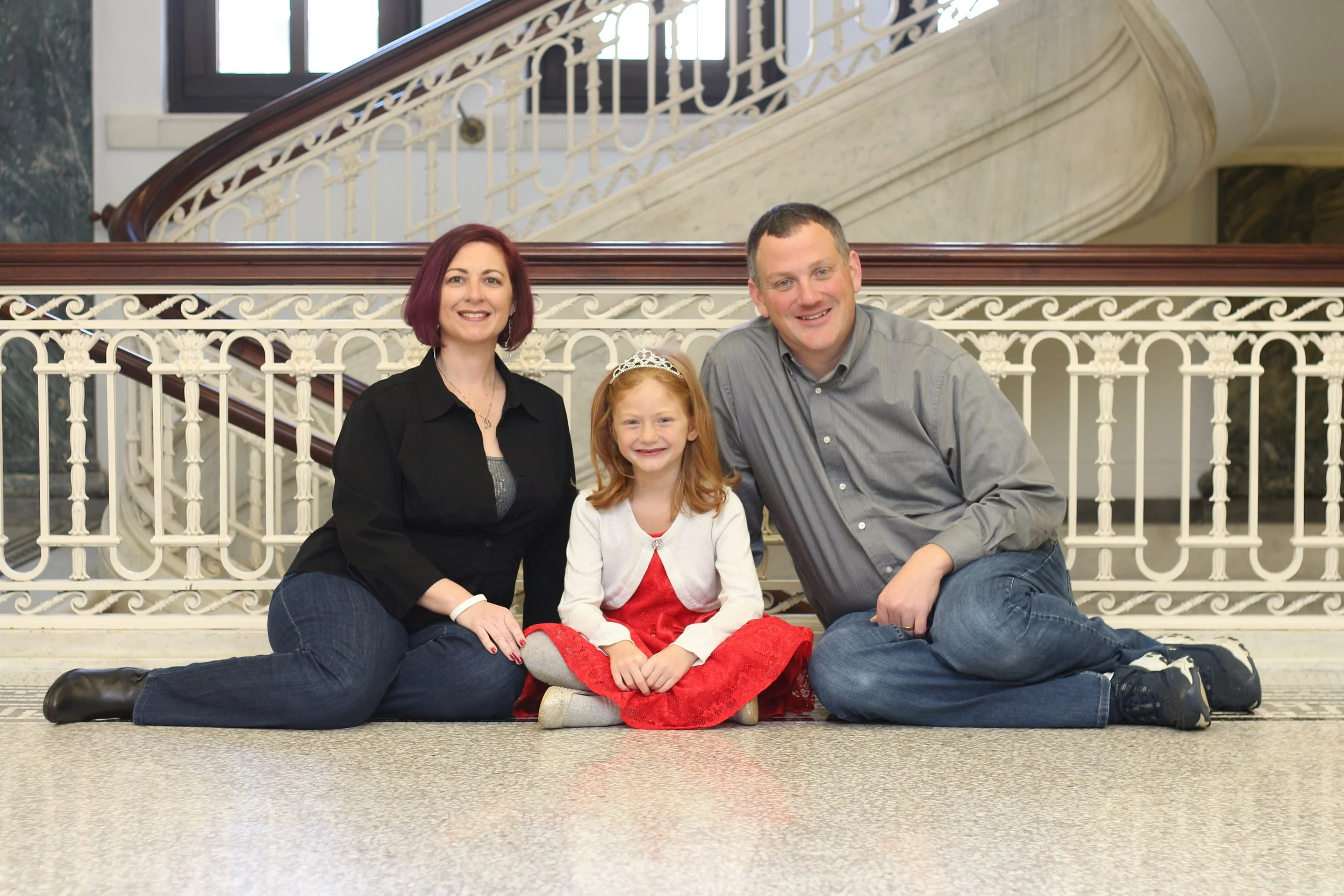A family of four sitting on the floor in front of a decorative staircase inside a building. The family includes a woman with red hair, a girl wearing a tiara and red dress, and a man with short hair in a gray shirt, all smiling at the camera.