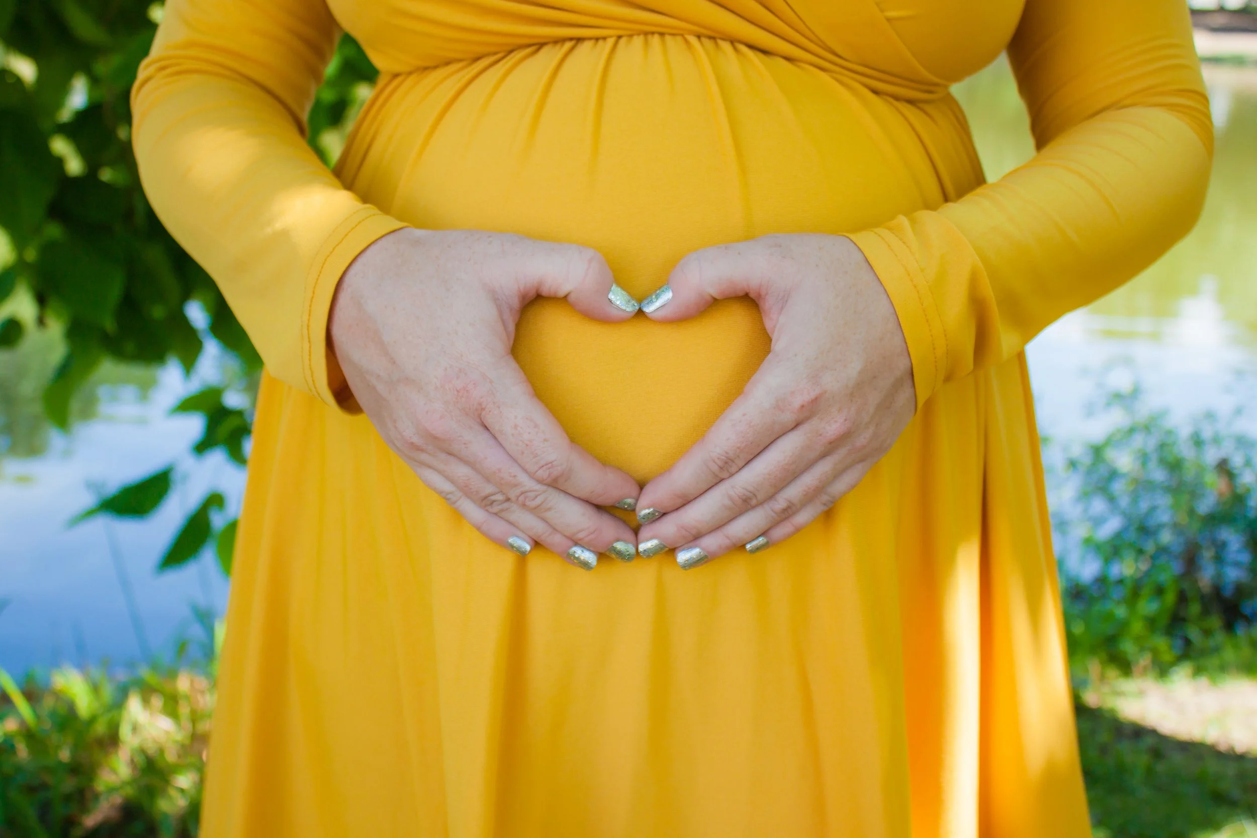 Person in a yellow dress forming a heart shape with hands over baby bump in an outdoor setting.