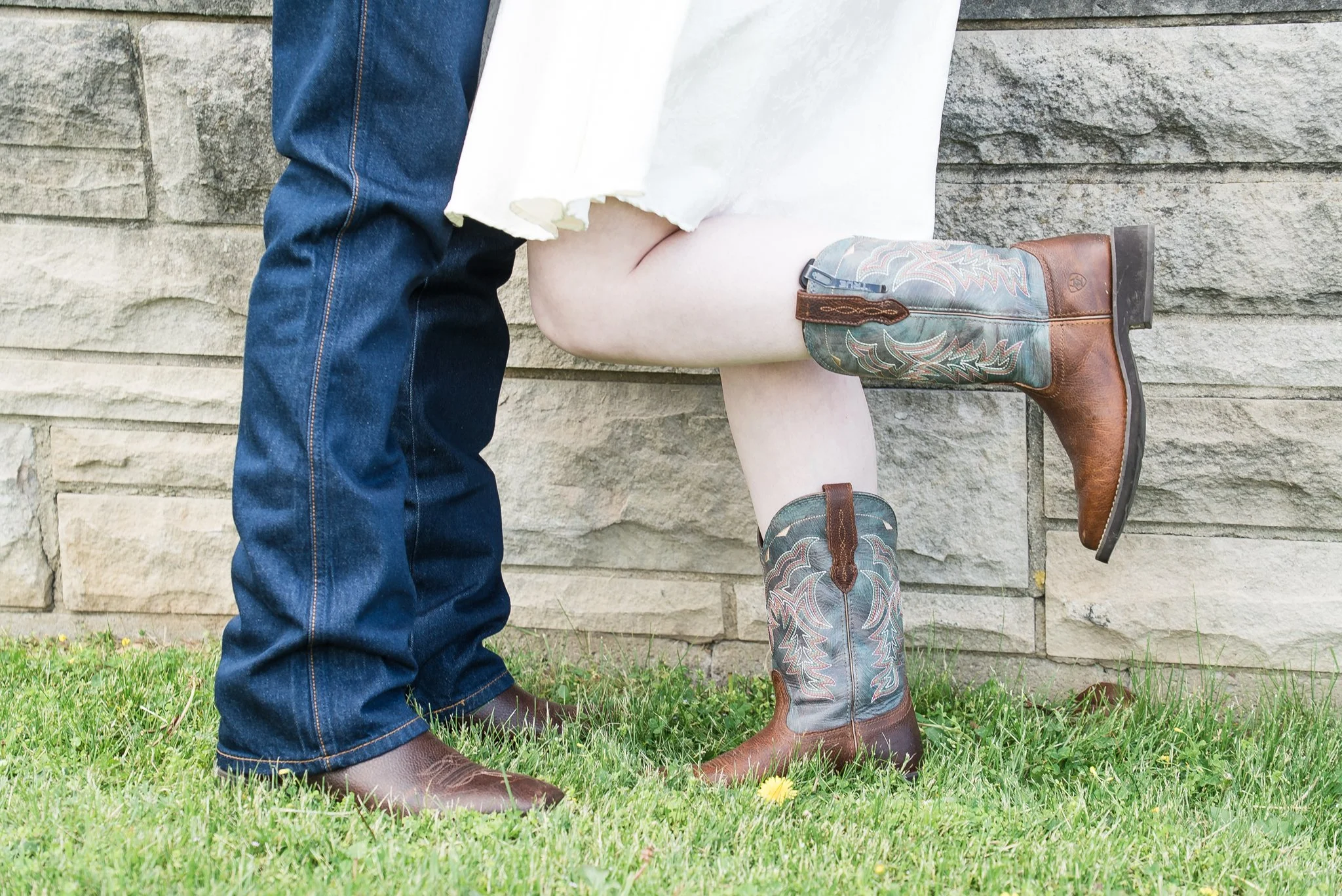 Close-up of two people, one wearing blue jeans and the other wearing cowboy boots, standing on grass with a stone wall in the background.