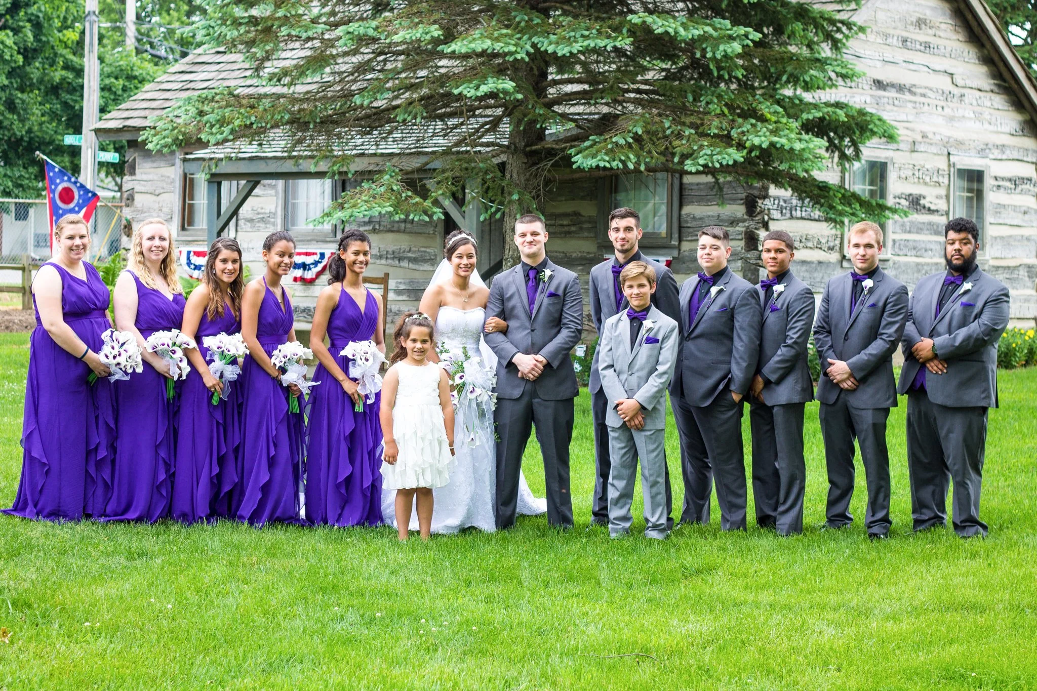 Group of people dressed in formal attire, including bridesmaids in purple dresses, a bride in white, and groomsmen in gray suits with purple ties, standing outdoors on a lawn with a rustic house in the background.