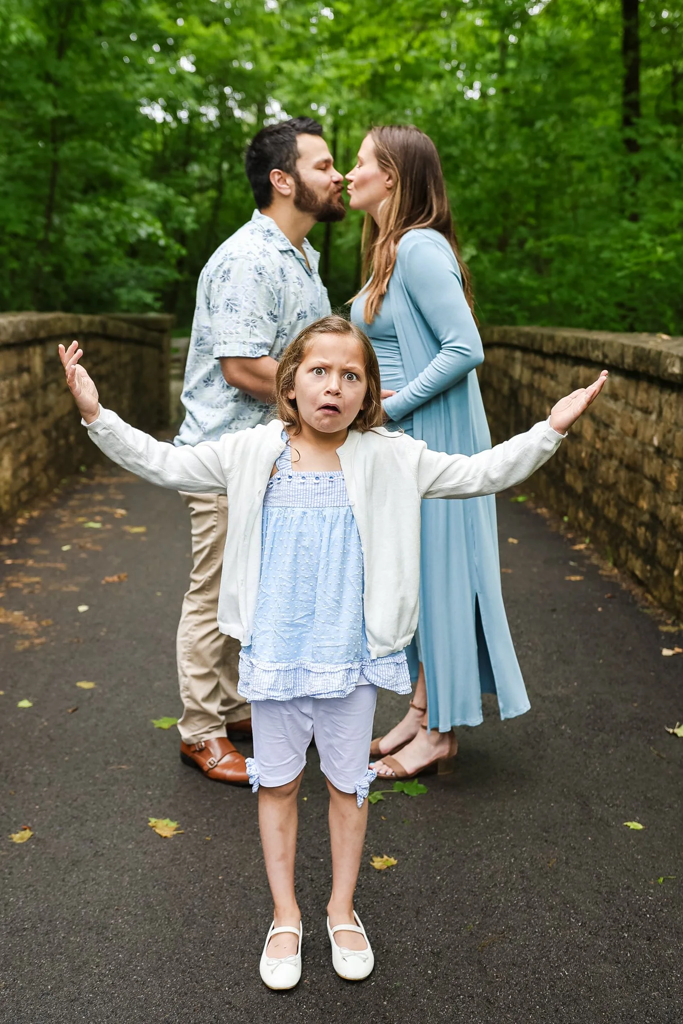 A young girl stands with her arms outstretched and a surprised expression as two adults, a man and a woman, in a wooded park lean in to kiss with closed eyes. The girl is wearing a light blue dress, white cardigan, and white shoes. The adults are dre