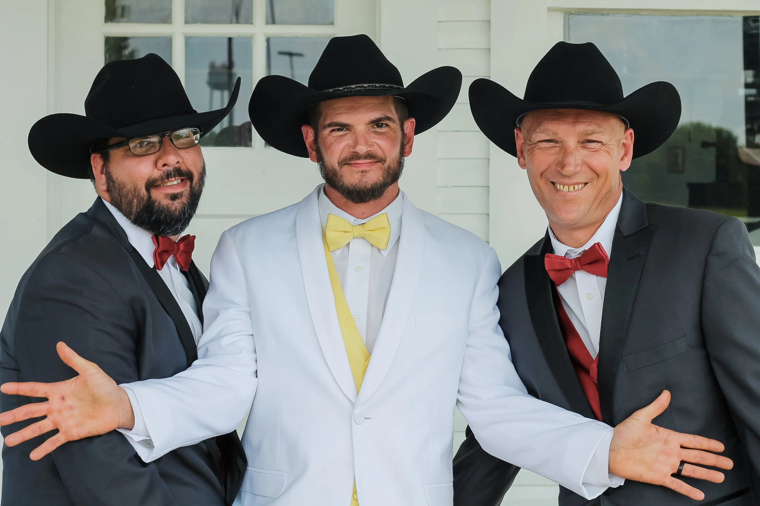 Three men dressed in tuxedos and bow ties, wearing large black cowboy hats, standing close together outside in front of a white house with windows.