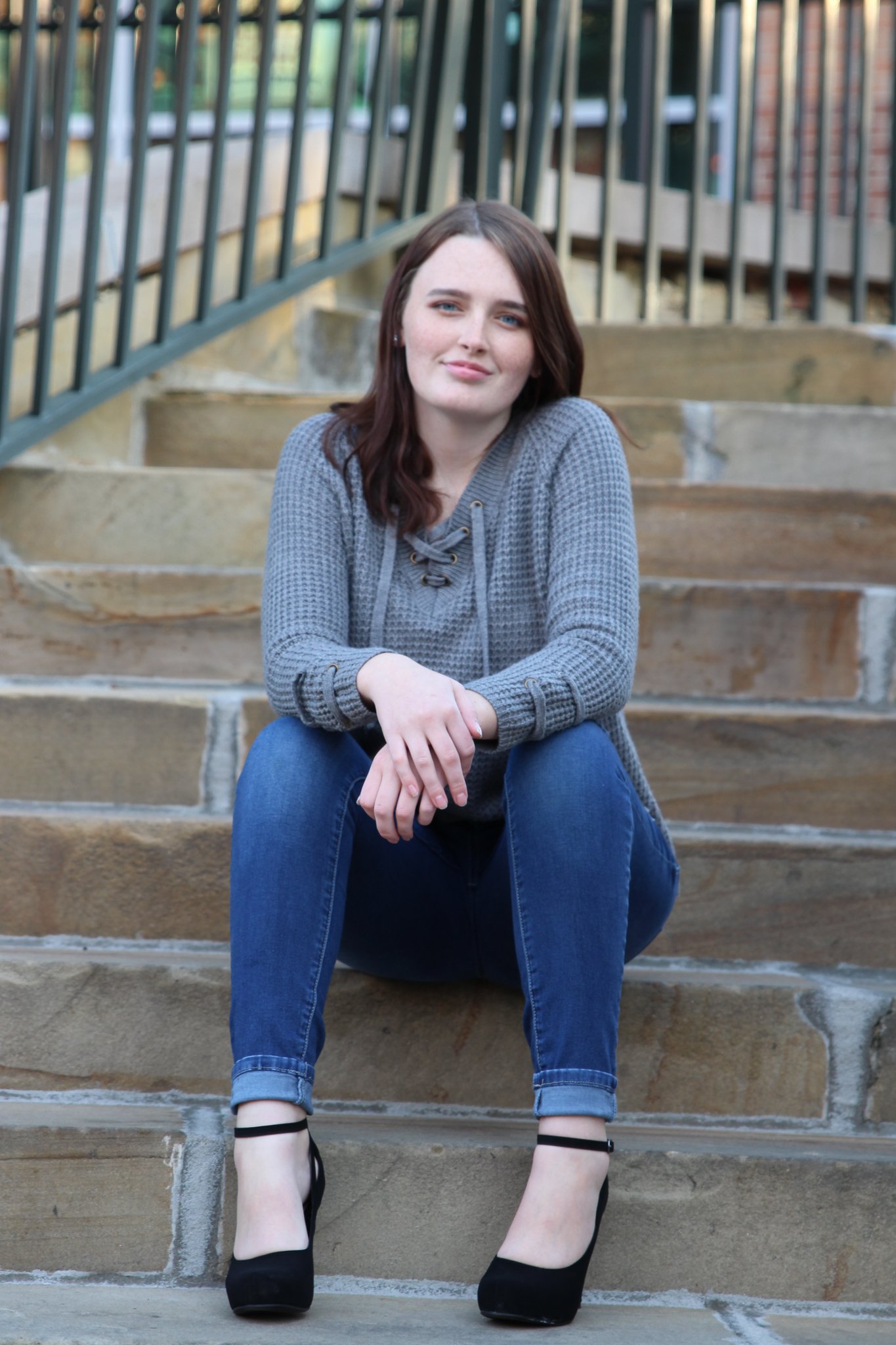 A young woman with brown hair and blue eyes sitting on outdoor stone steps with black railings, wearing a gray sweater, blue jeans, and black high heels, looking at the camera.