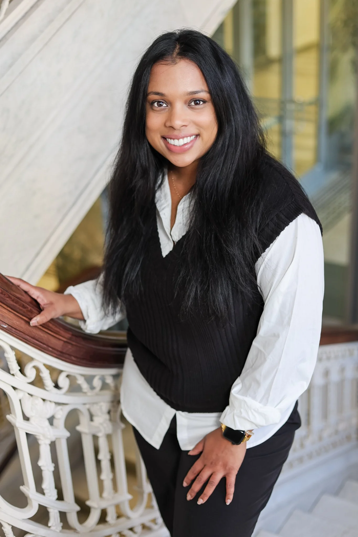 A woman with long black hair wearing a white blouse and black vest, standing near a decorative white railing with a wooden handrail, smiling at the camera.