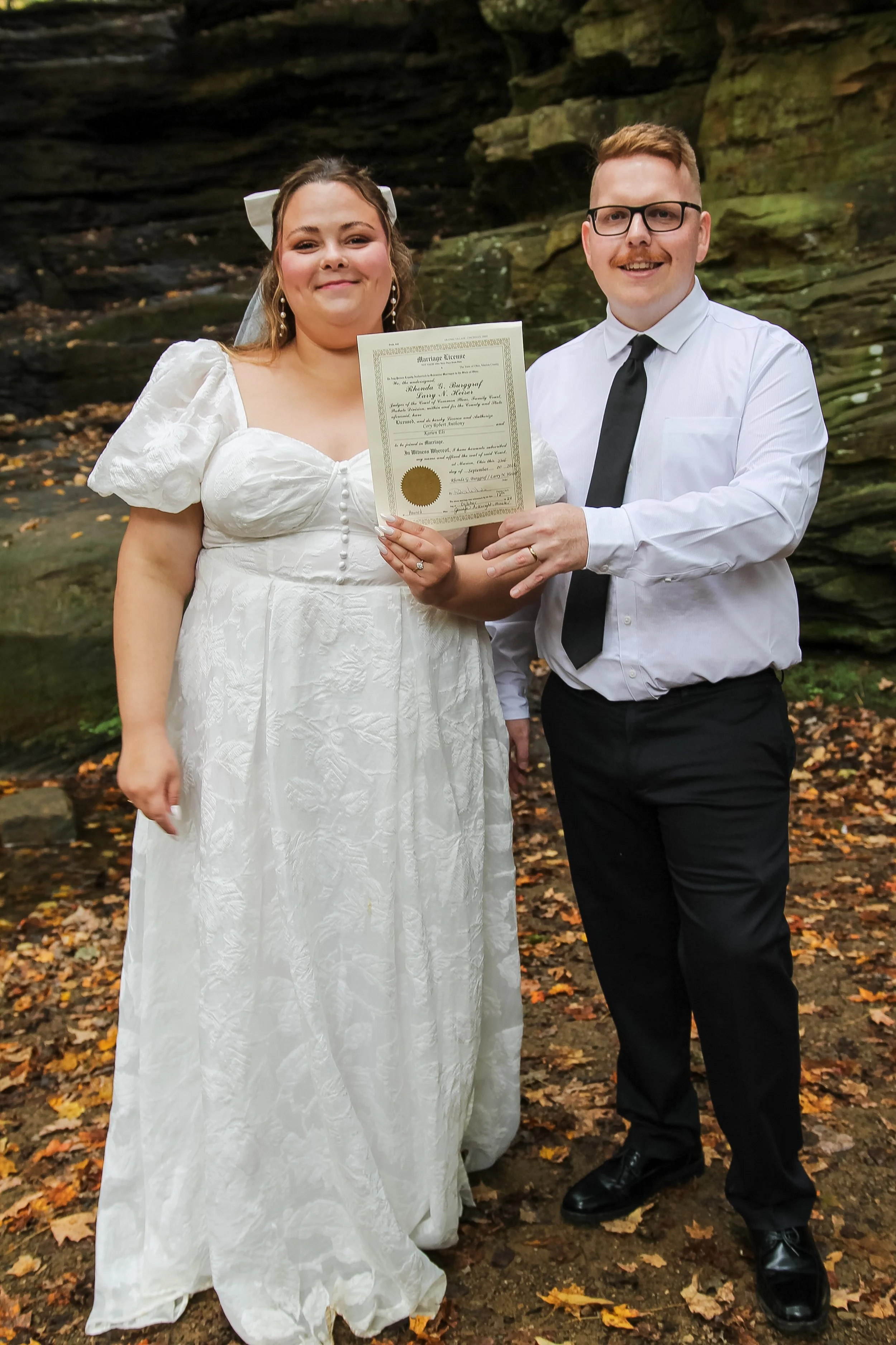 A bride in a white wedding dress holding a marriage certificate, standing next to a groom in a white shirt, black tie, and black pants, outdoors with fallen autumn leaves and rock formations in the background.