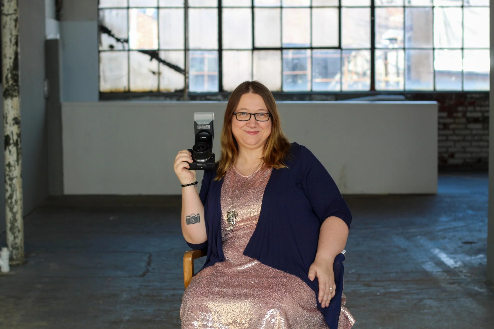 A woman with glasses and light brown hair sitting in a chair inside an industrial-style building with large windows, holding a camera in her right hand, smiling at the camera.